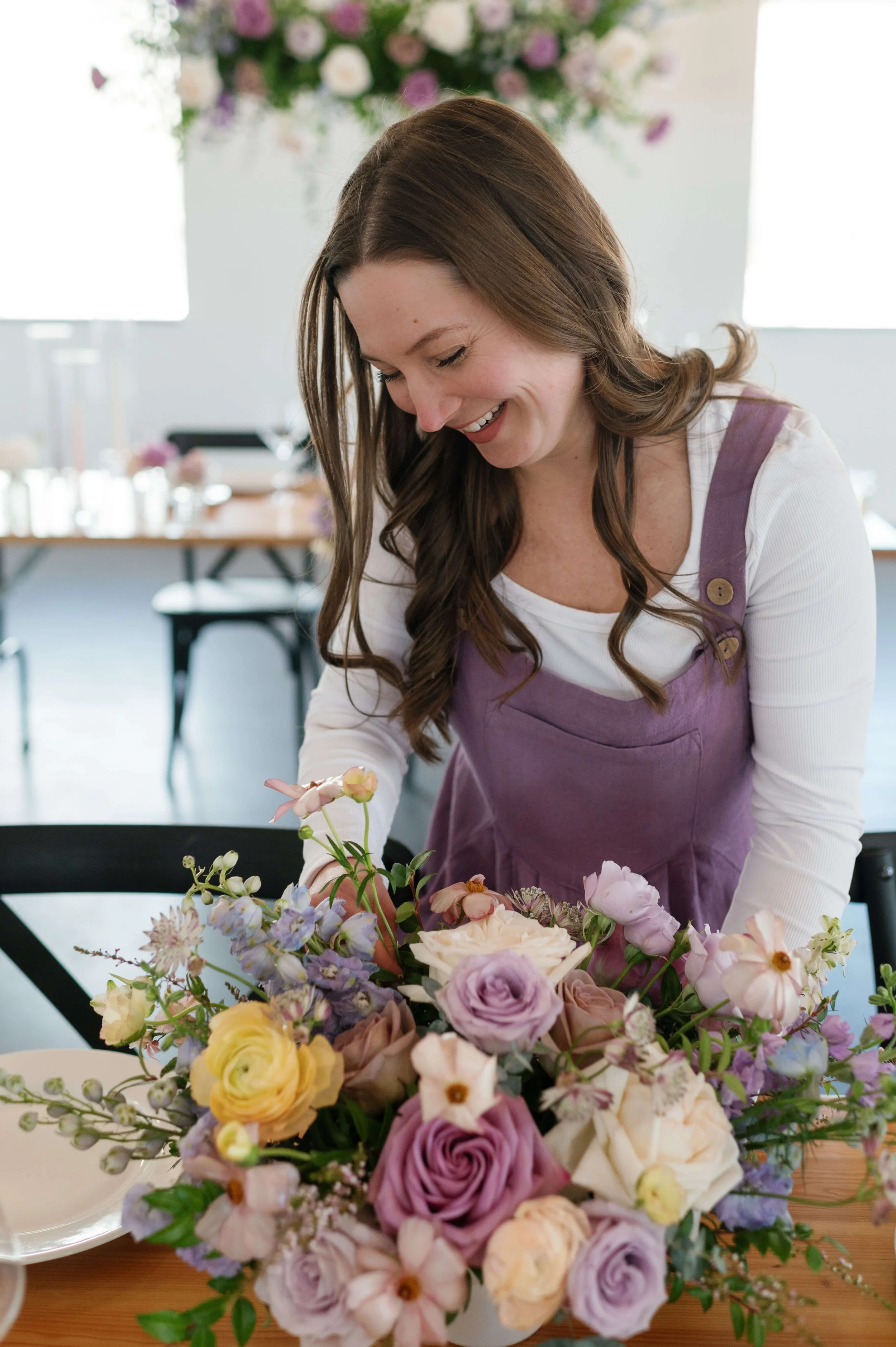 Beth, florist and owner of For Floral Sake, smiling while creating a floral arrangement at her work table.