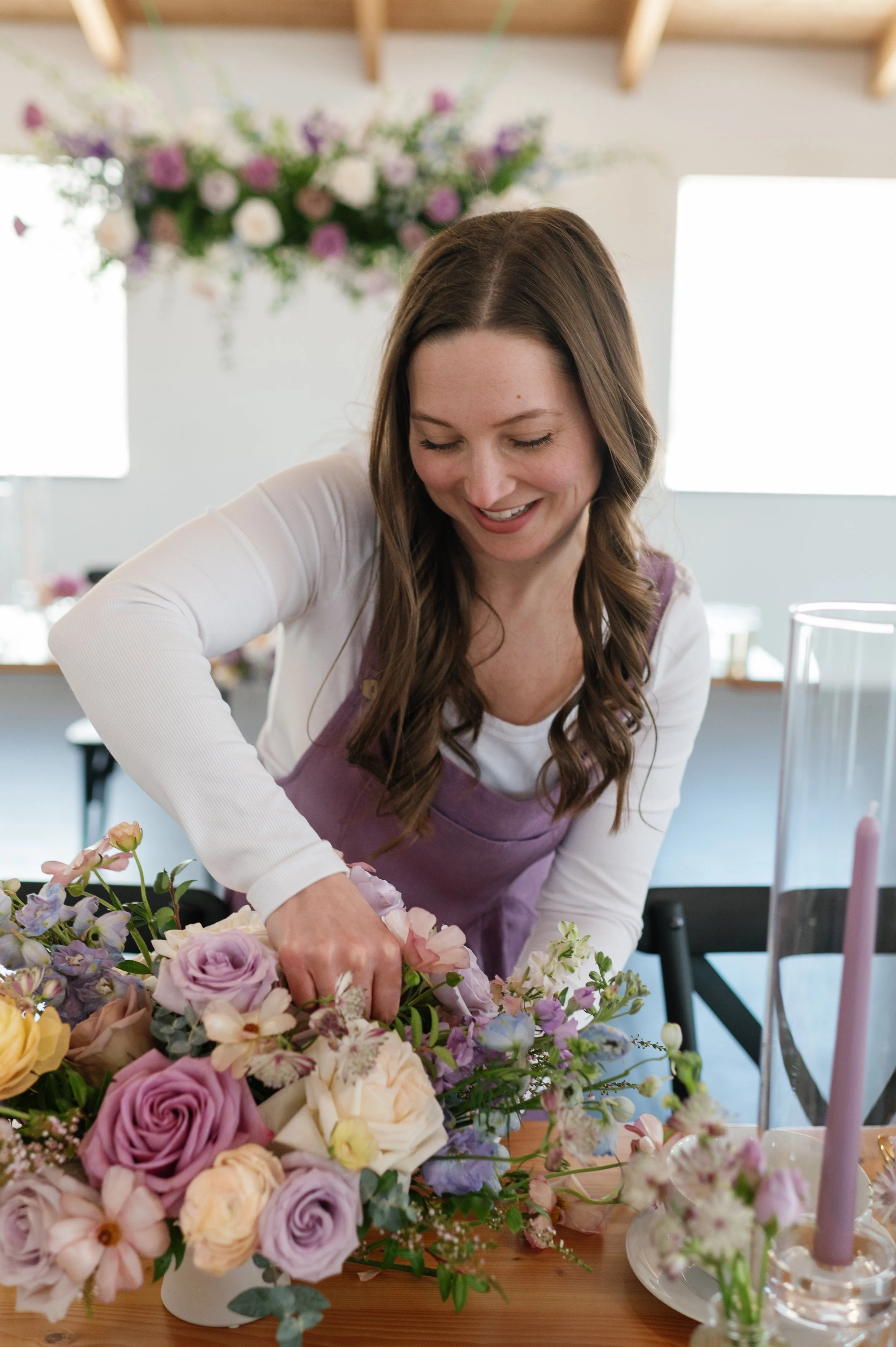 Smiling luxury wedding florist in Duluth, MN creating a lush floral centerpiece beneath a hanging flower installation in soft purple and blush tones.