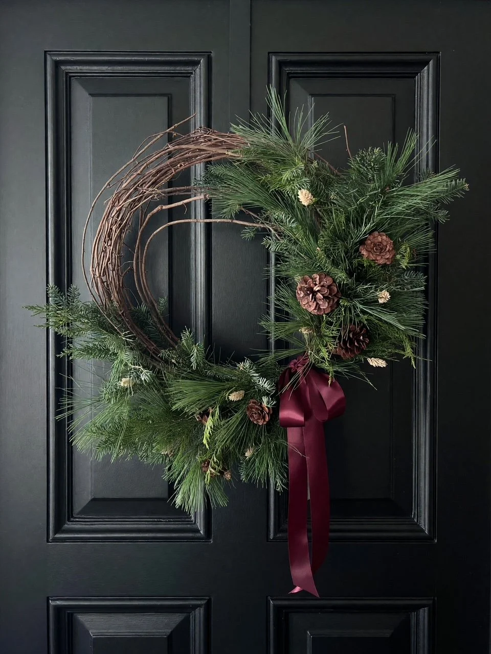 Holiday grapevine wreath with winter evergreens, pinecones, dried accents, and satin ribbon on a black door