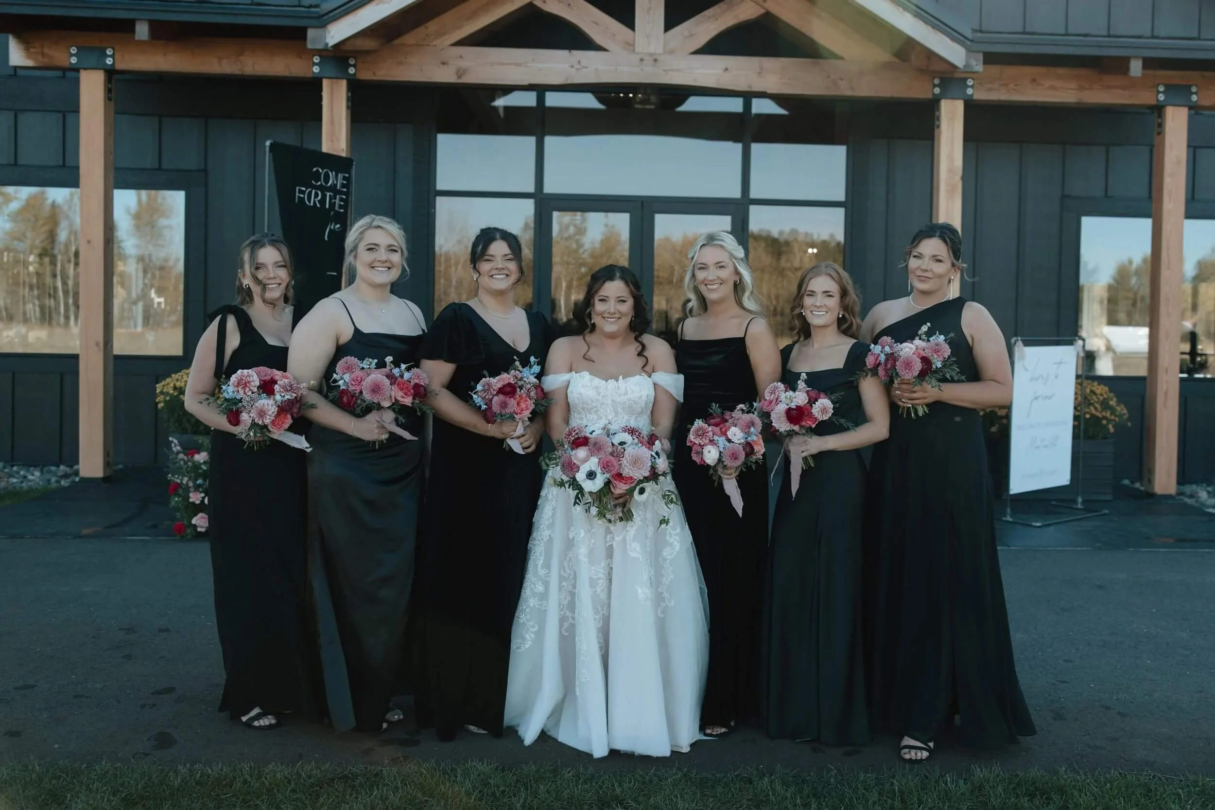 Bride and bridesmaids holding colorful bouquets in front of the black exterior of Ivy Black in Duluth, Minnesota.