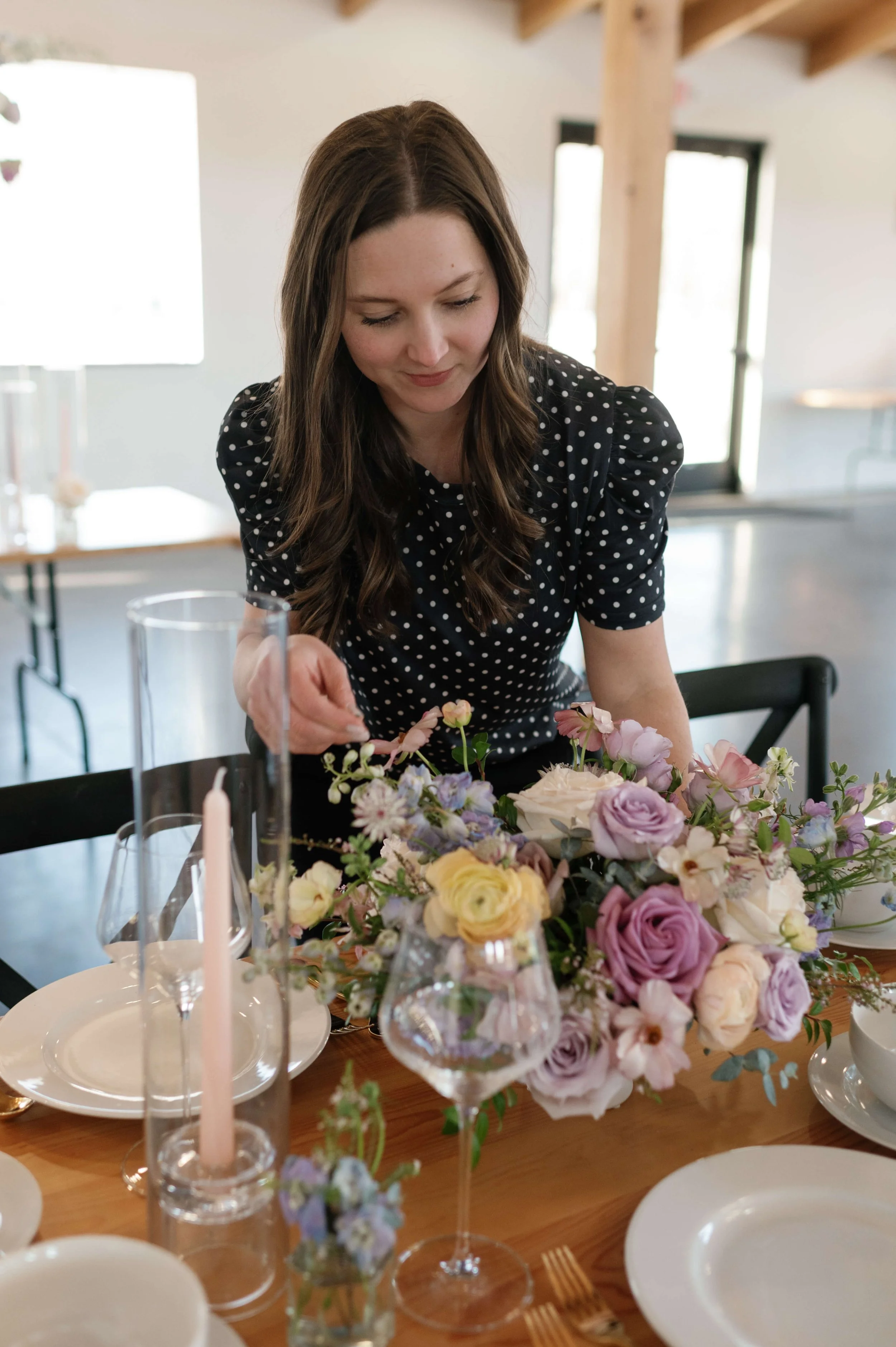 Woman in black polka dot dress arranging a floral centerpiece on a dining table set with plates, glasses, and candles.