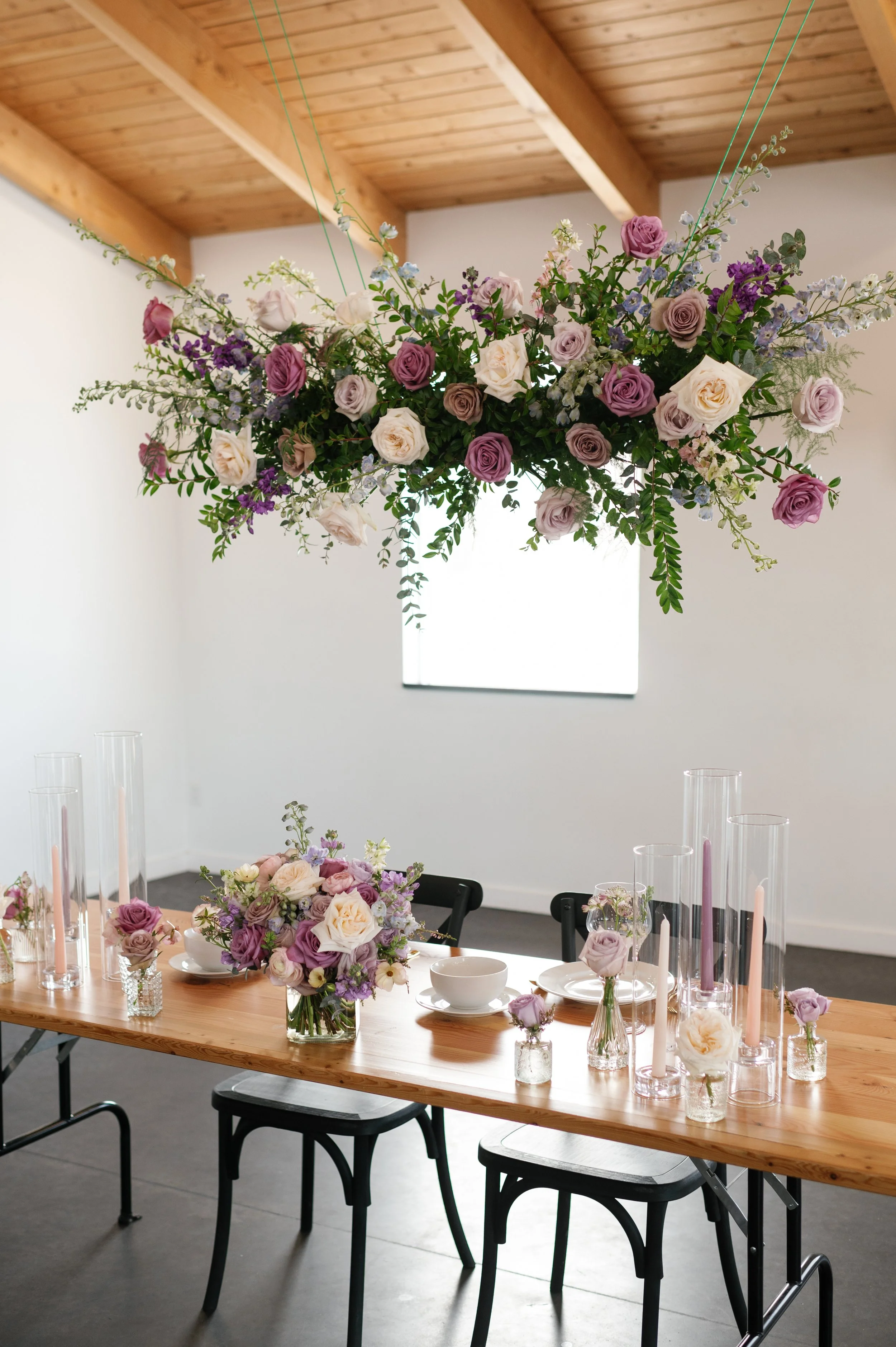 A dramatic hanging floral installation paired with lush tabletop blooms, candles, and bud vases in soft purples and blush. Created by a full-service wedding florist in Duluth, MN, this luxury floral design brings modern elegance and atmosphere to wed