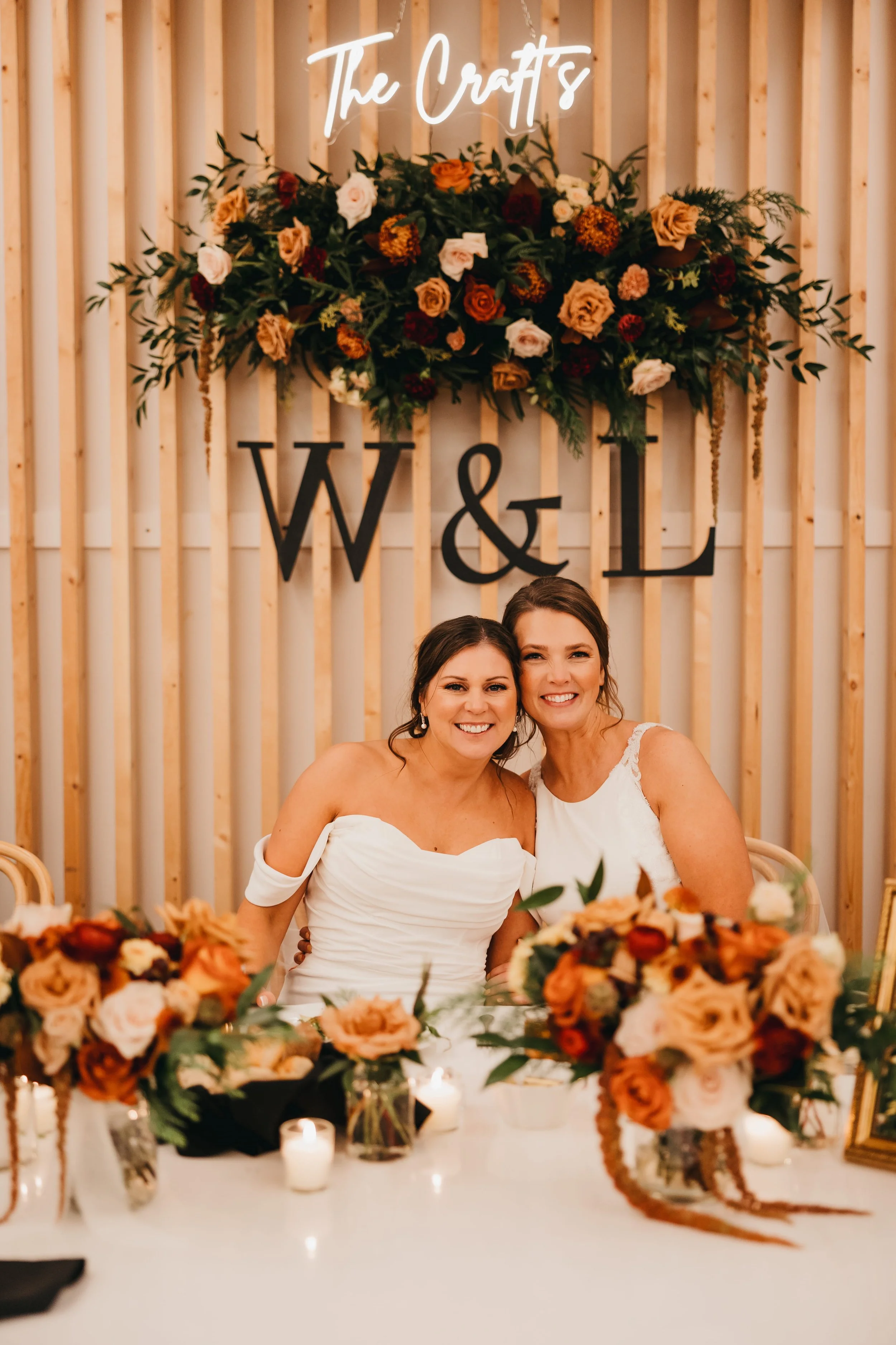 Two brides seated at the head table with florals and candles at a Nisswa wedding flowers designed by For Floral Sake