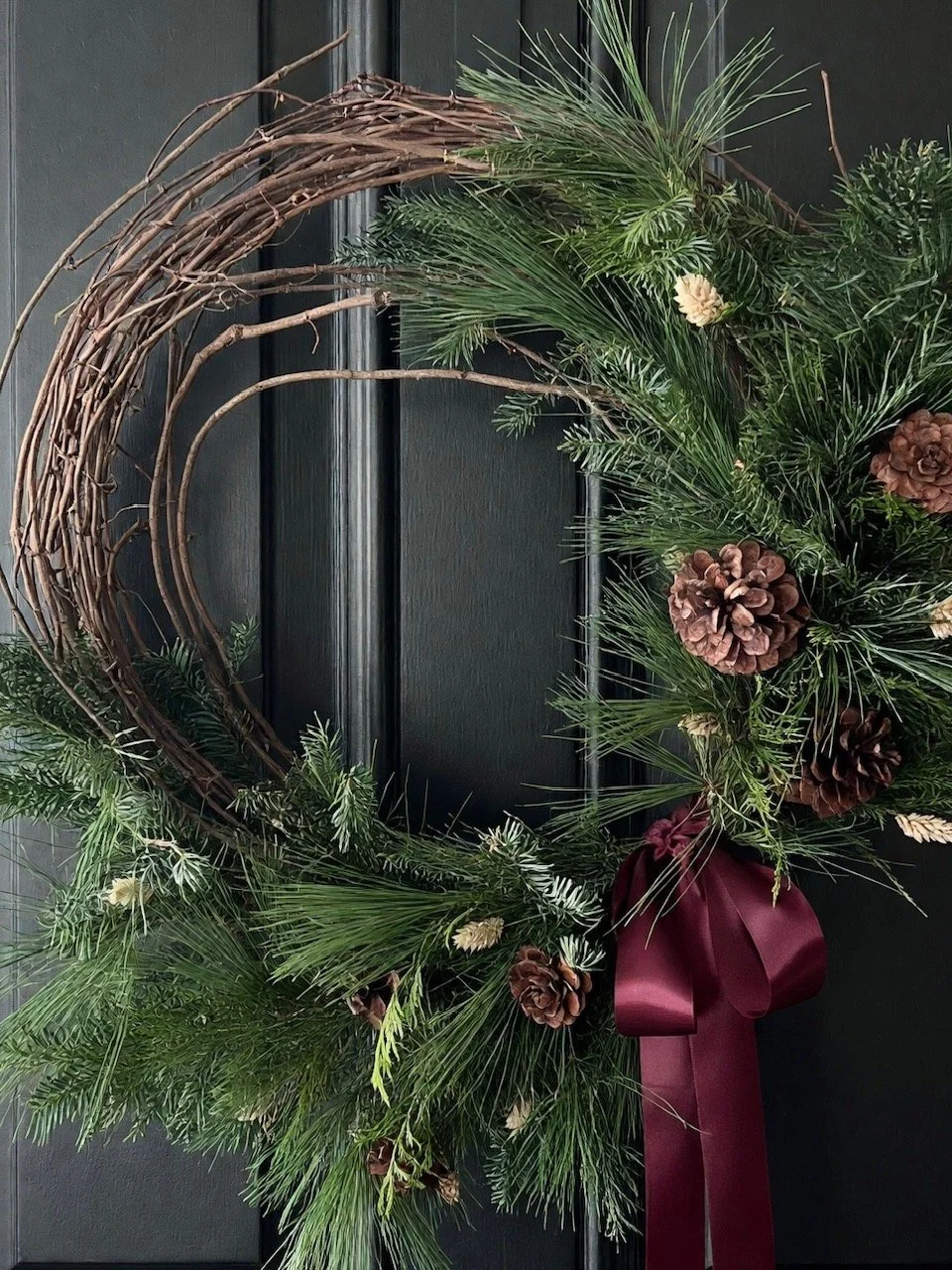 Close-up of a winter wreath with pine, cedar, spruce, pinecones, dried accents, and satin ribbon