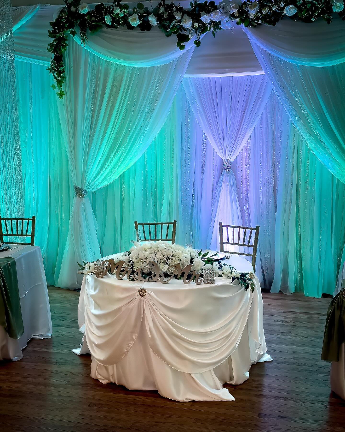 Soft ivory draping, luminous turquoise lighting, and gold Chiavari chairs frame the sweetheart table. White roses complete the timeless&nbsp;&nbsp;setting.#cherryhillpark #marylandweddingvenue #backyardwedding #marylandbride #dmvweddings #mdweddingve