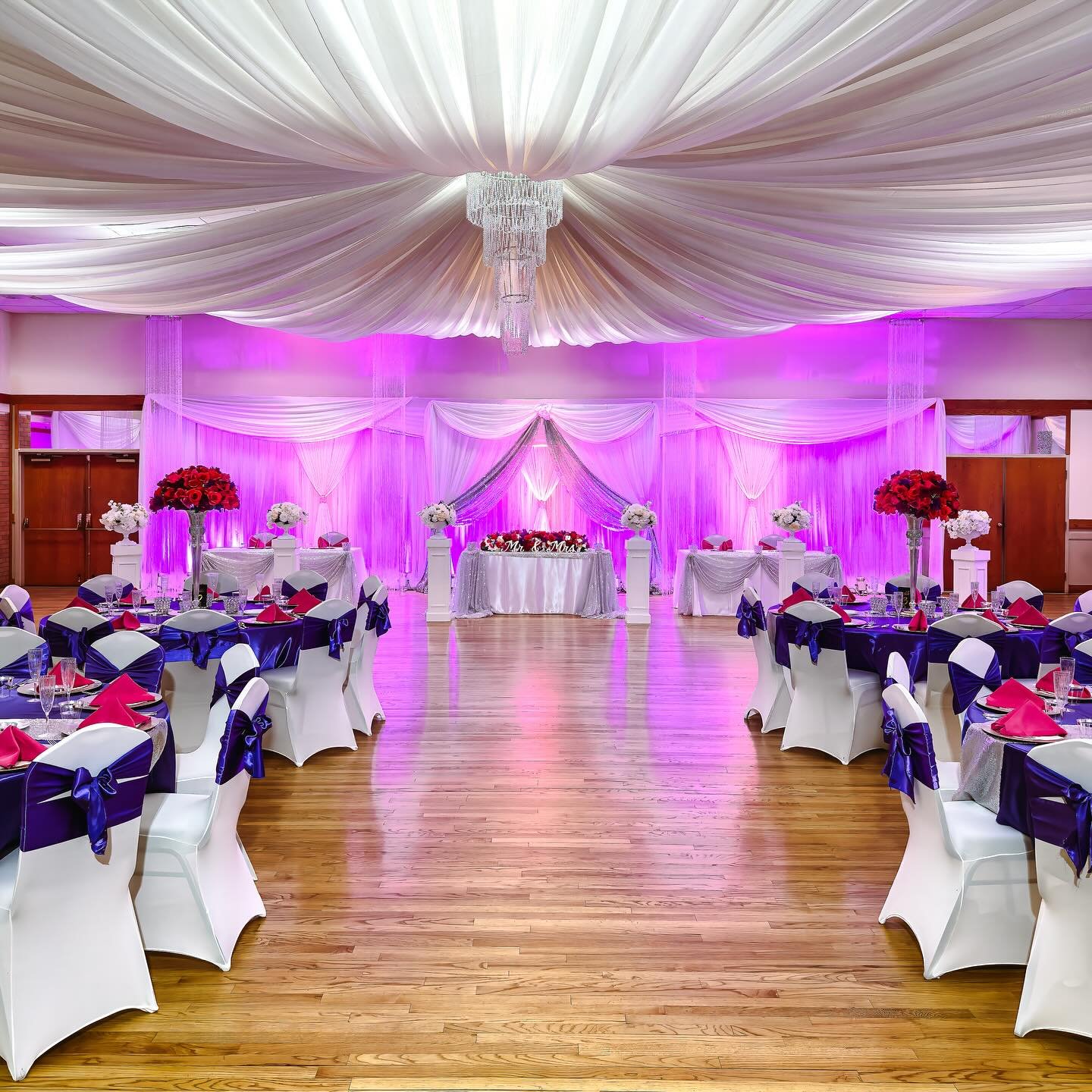 Unusual floor plan with guests seated at tables during the ceremony and sweetheart table moved into ceremony spot during cocktail hour. Lush centerpieces bloom in shades of crimson and ultraviolet, while lilac uplighting washes over the sheer white d
