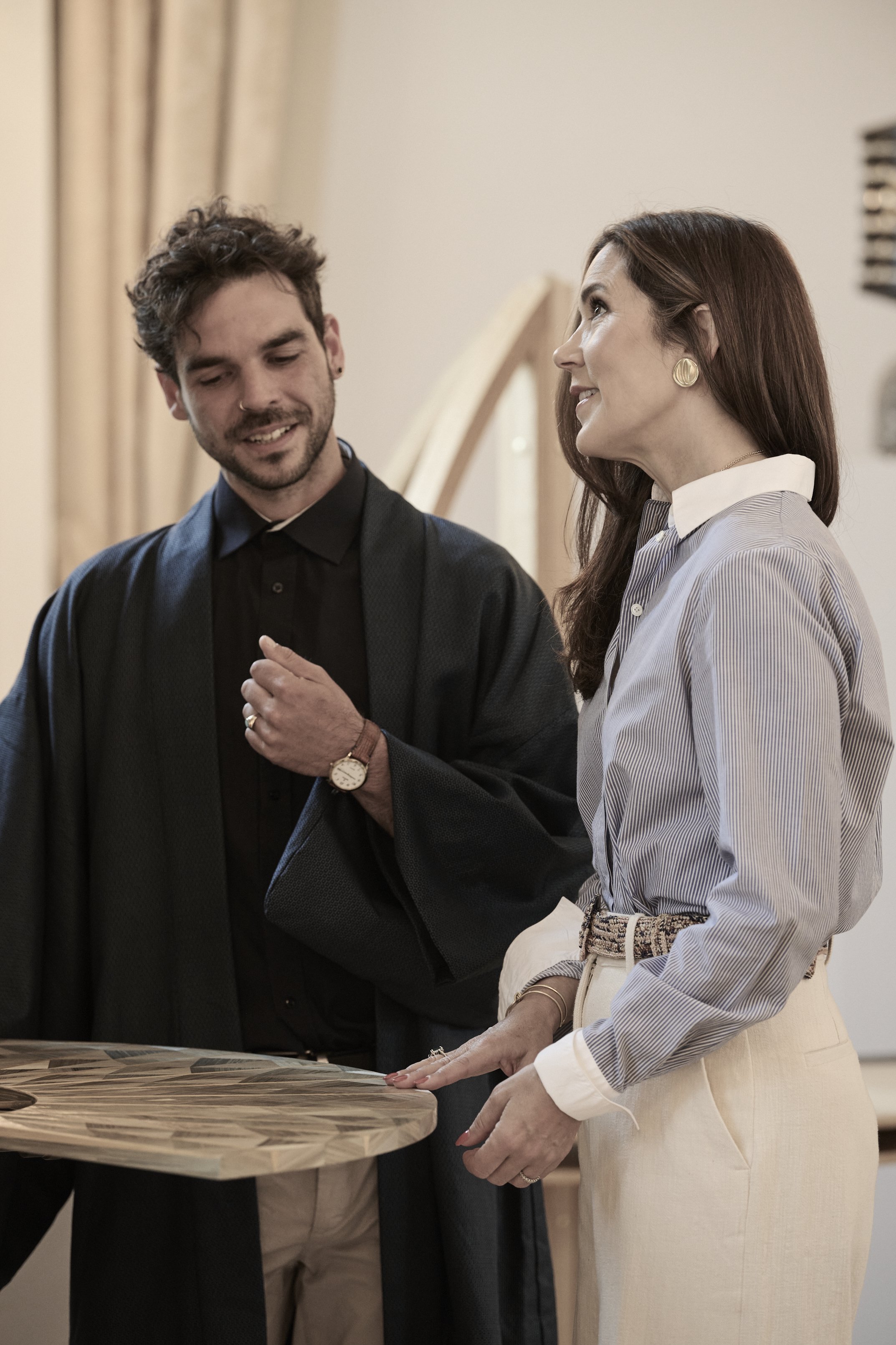 A man and a woman are standing together, smiling and talking. The man has short, curly hair, a beard, and is wearing a black jacket over a black shirt. The woman has long, brown hair, and is wearing a light striped shirt with large gold earrings and 