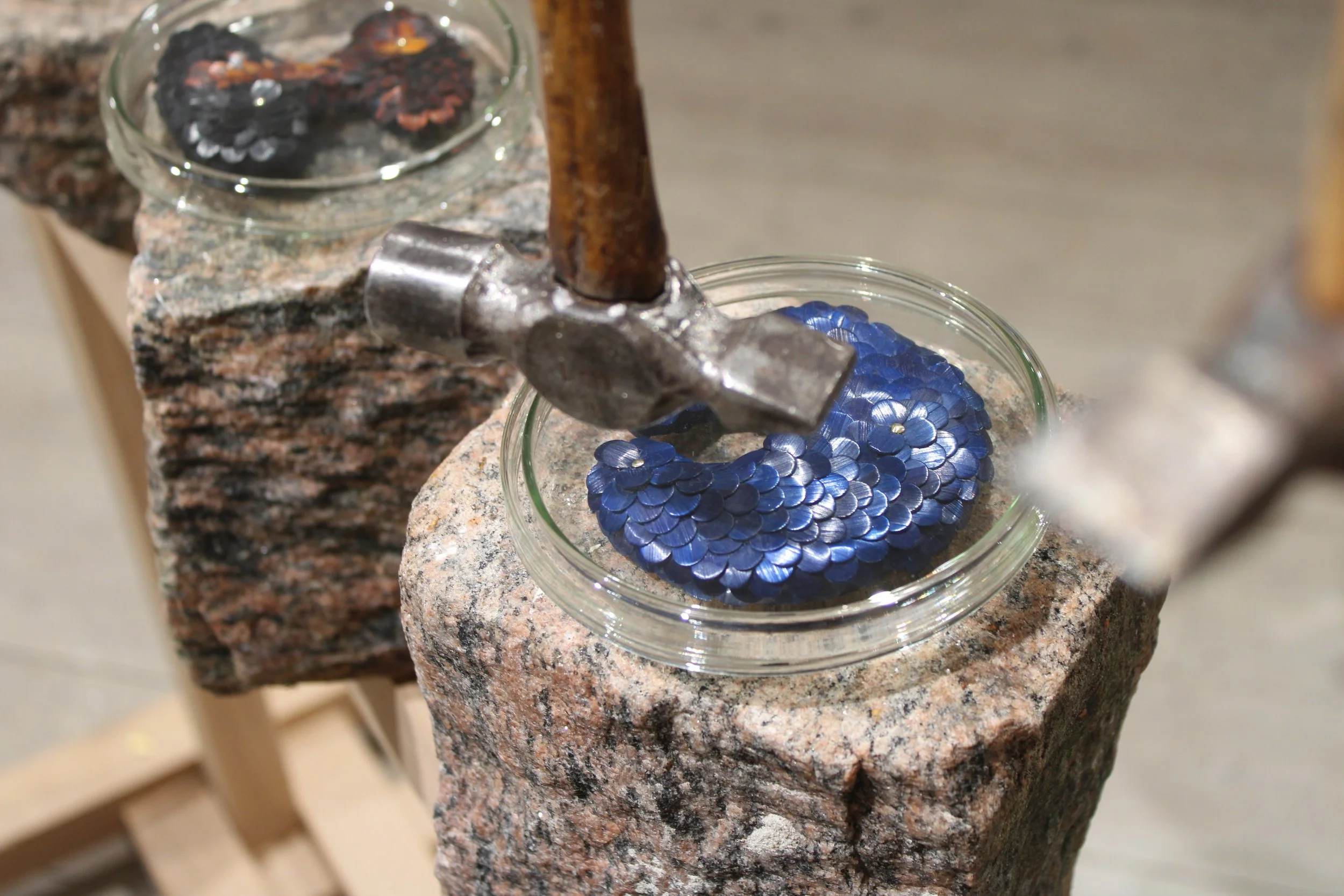 Invisible monster 22 by Jonathän Jouan Studios. A glass jar filled with small blue flower-shaped sequins placed on a granite countertop. In the background, part of a hammer and another jar with black and reddish-brown objects are visible.