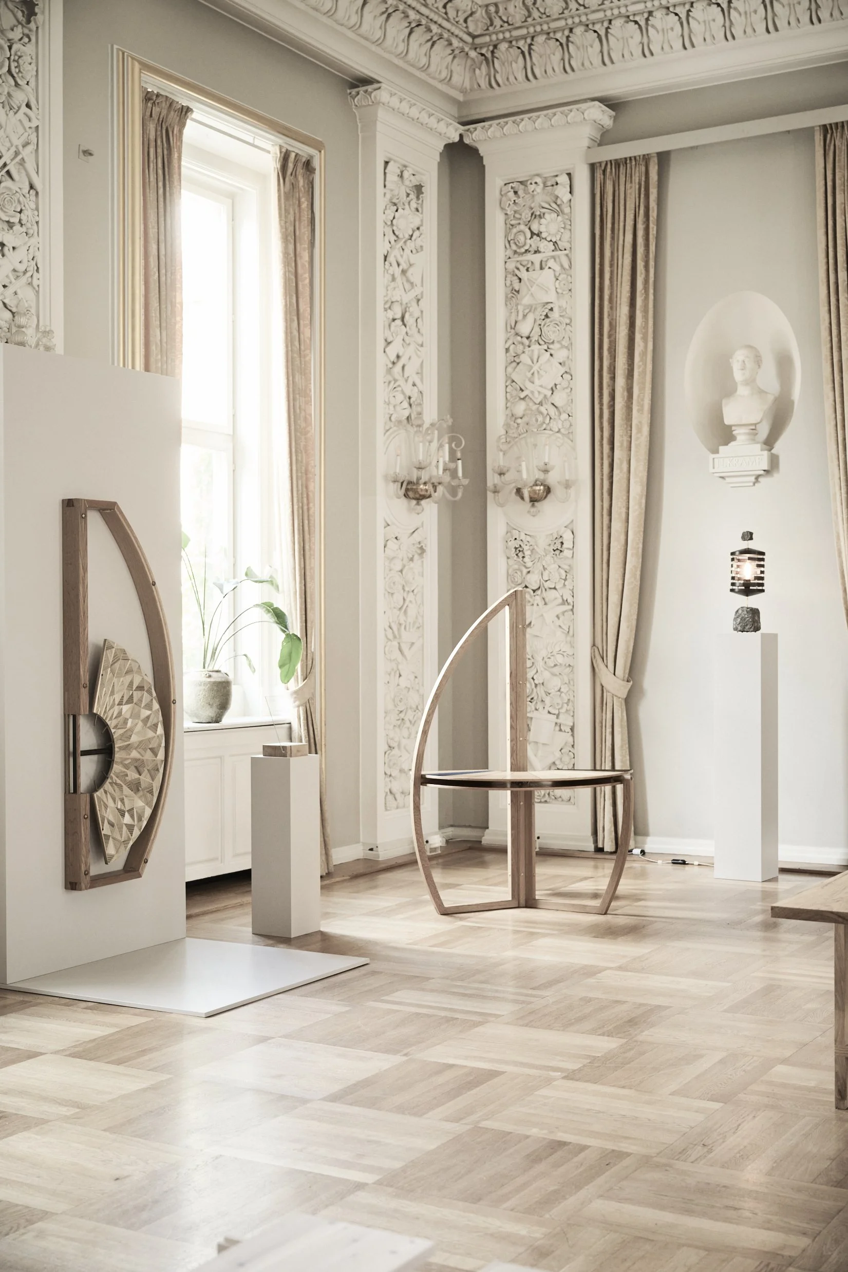 Interior of a room with ornate white plaster moldings, a window with curtains, and a decorative bust on a pedestal. There are wooden furniture pieces and a wooden floor.