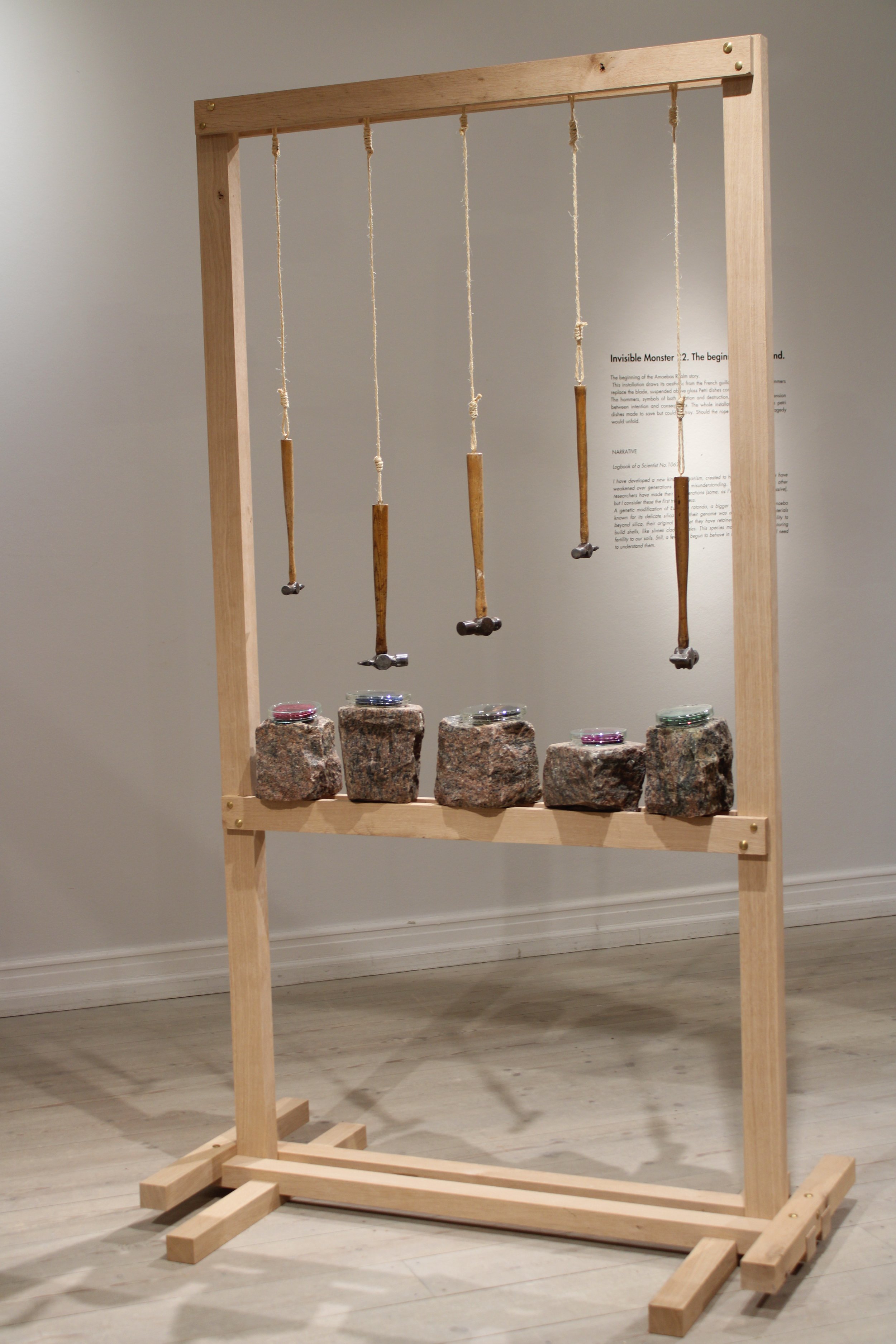 Invisible monster 22 by Jonathän Jouan Studios of the Amoebas Realm. An art installation features a wooden frame with five hanging hammer objects made of wood and string above five stone blocks with small petri dishes on top, in a gallery setting.