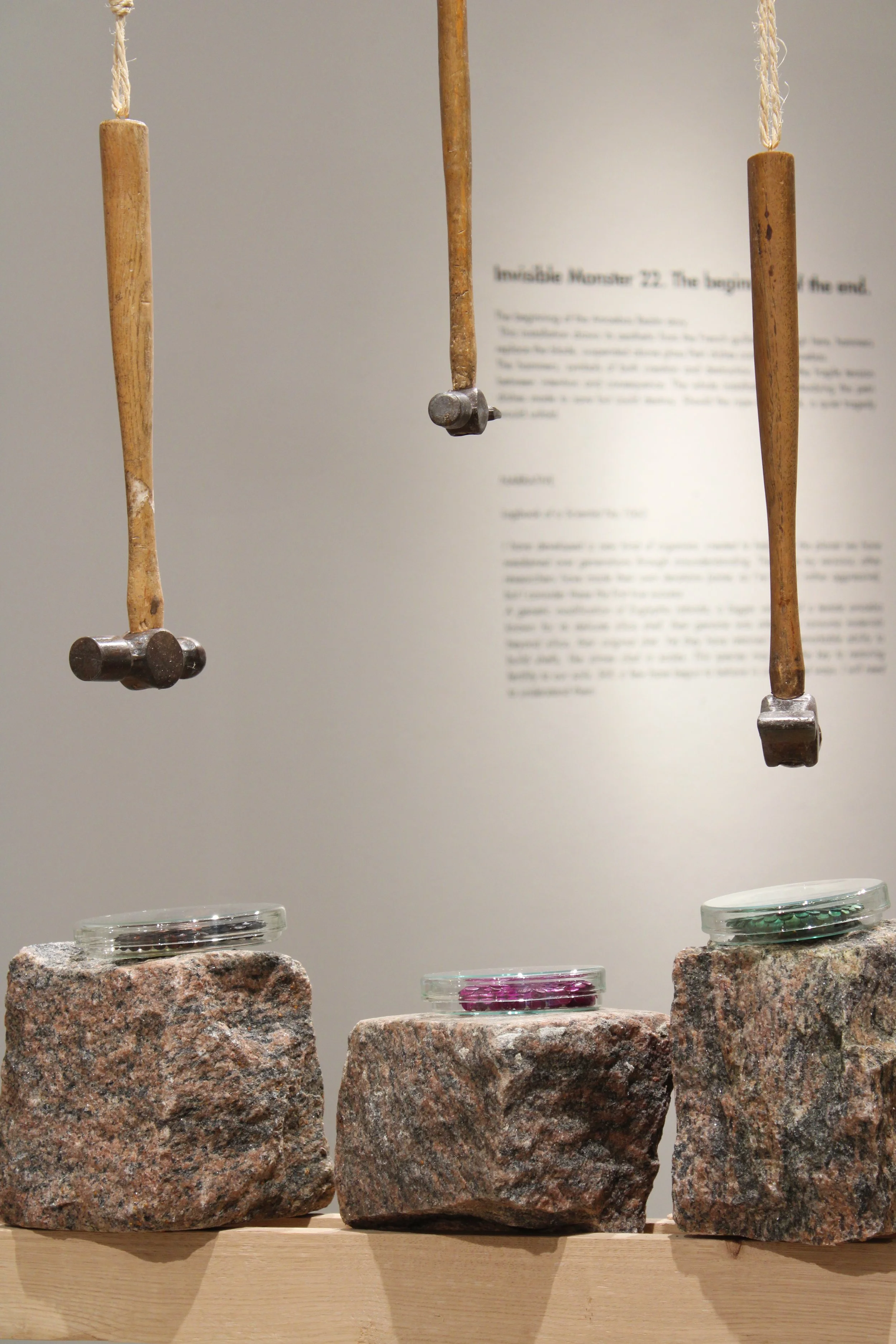 Three wooden hammers hanging from strings, displayed above a row of large granite blocks, with small glass dishes filled with colorful glass pieces on top of the stones.