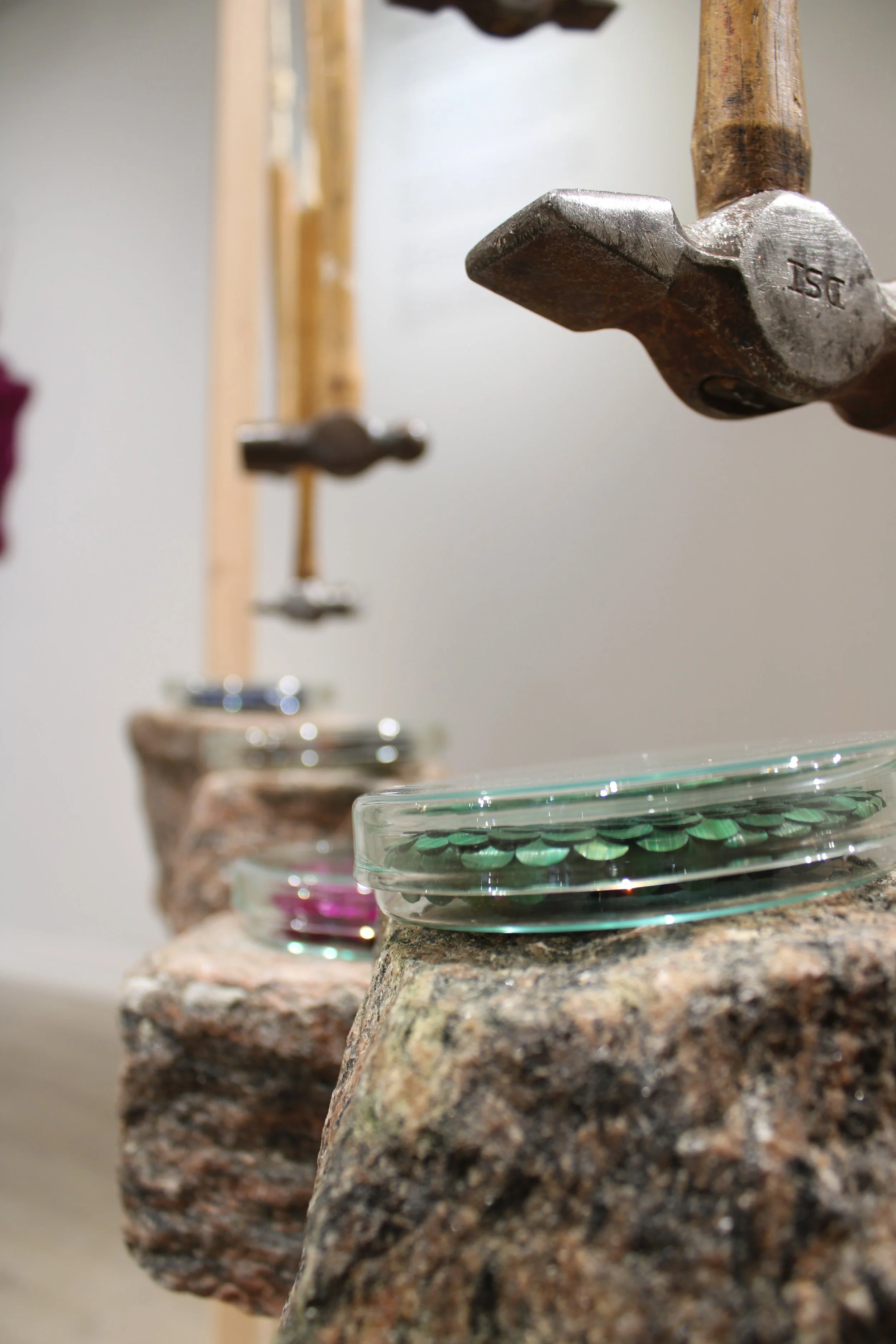 Close-up view of a row of small glass bowls on a irregularly shaped, multicolored stone surface, with a focus on a metal axe mounted on the stone. The bowls contain colorful decorative items, possibly beads or stones.
