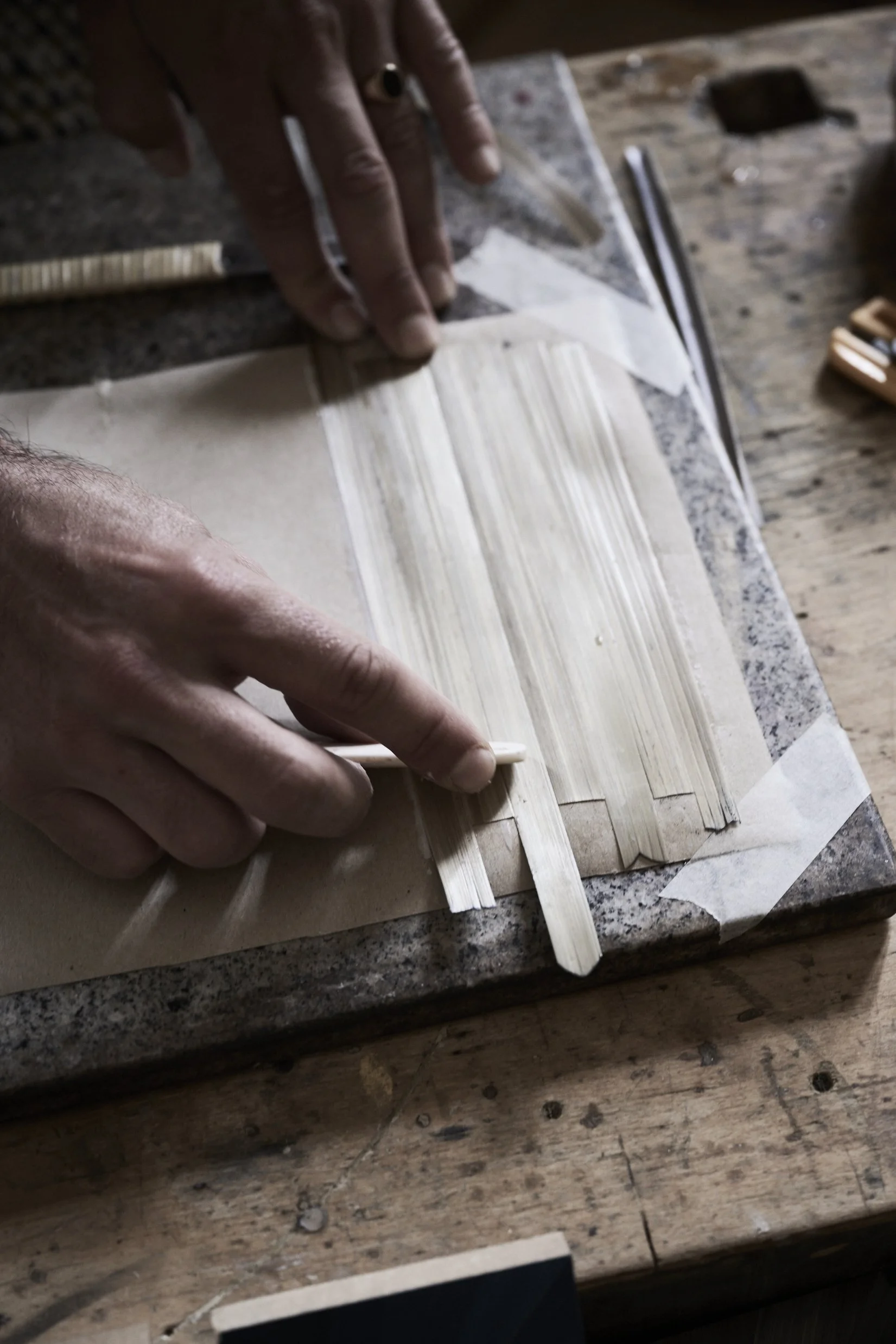 Close-up of hands working with thin wooden strips placed on a piece of paper or thin surface, on a wooden workbench with tools nearby.