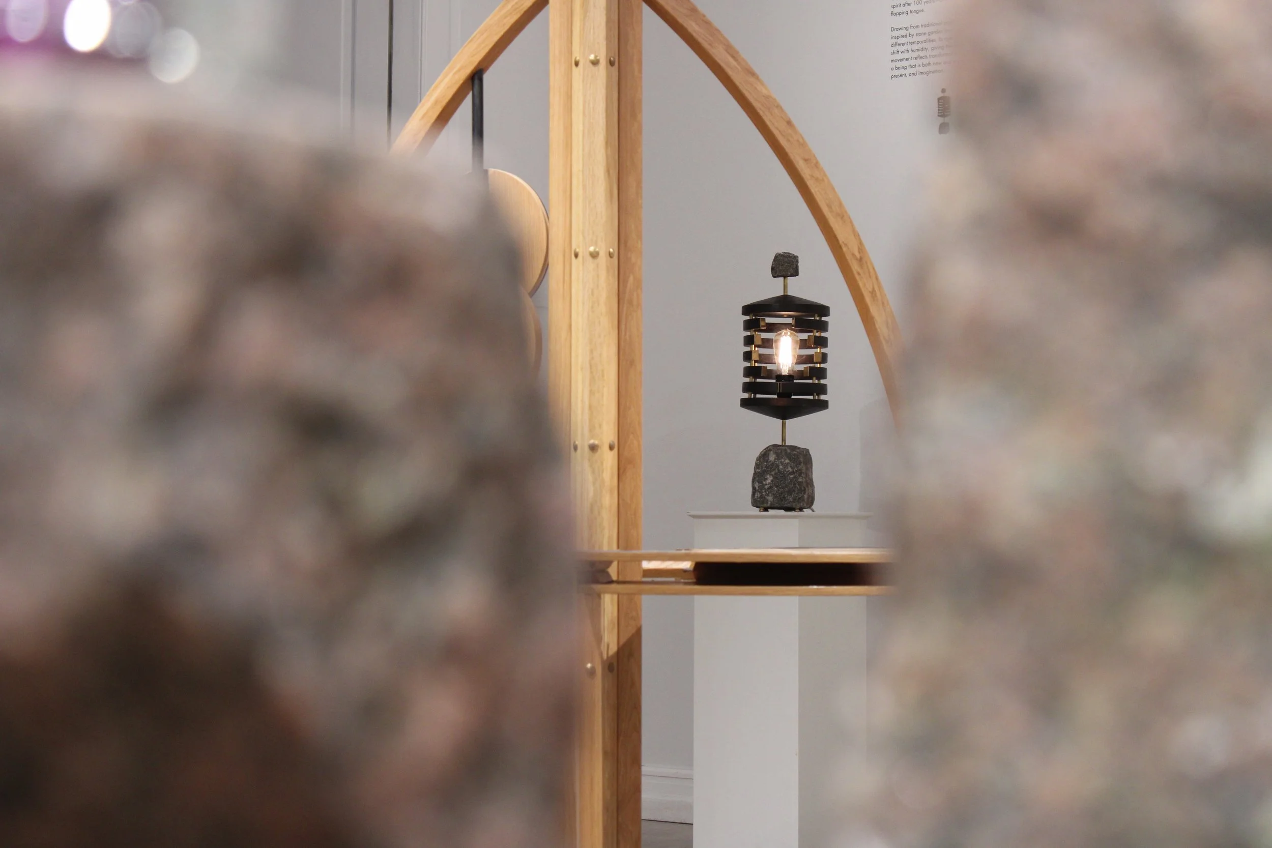 An art installation featuring a black lantern with a visible light bulb, mounted on a stone base, placed on a display table with a wooden frame in the background. Blurred objects in the foreground partially obscure the view.
