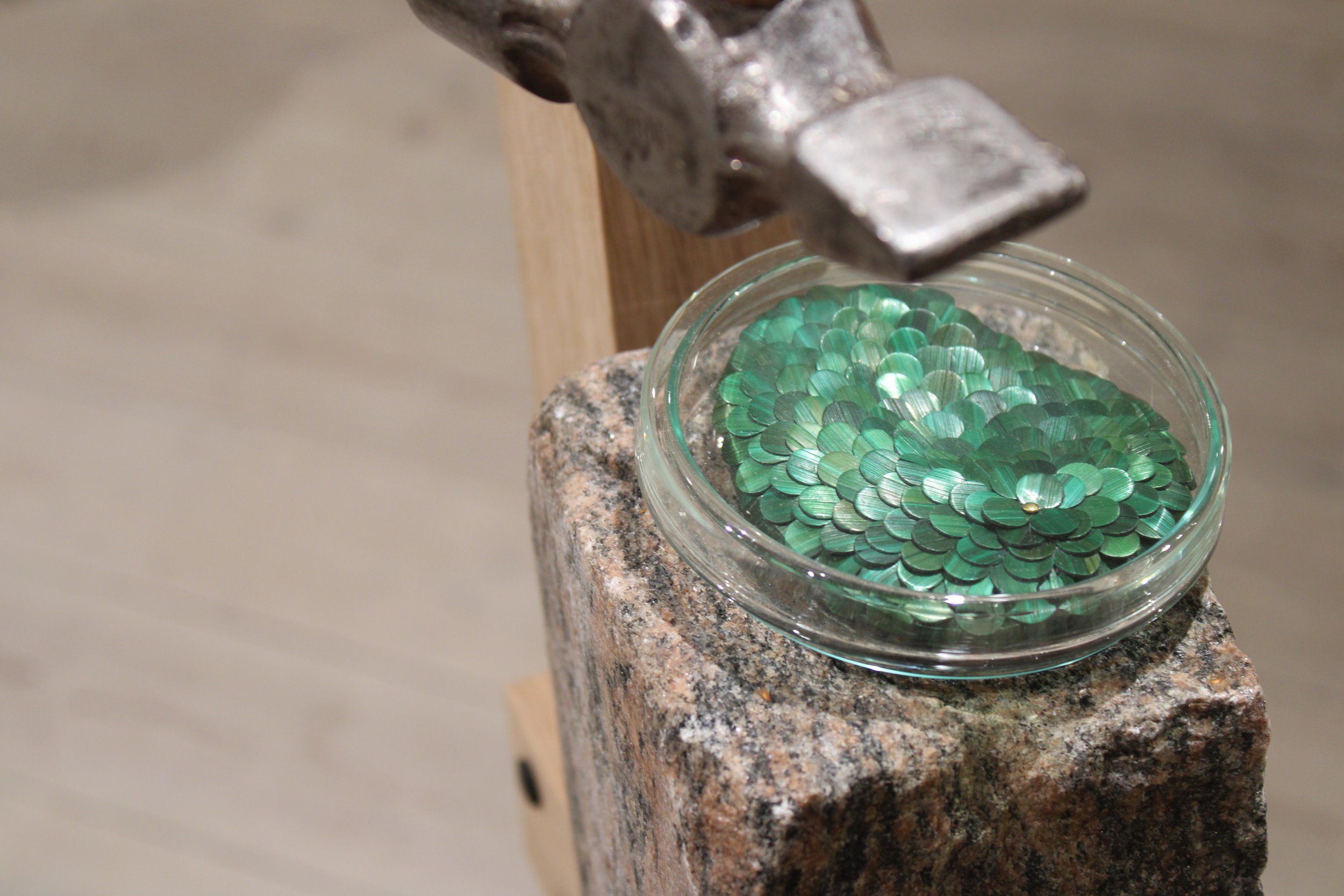 Invisible monster 22 by Jonathän Jouan Studios of the Amoebas Realm collection. A close-up of a glass bowl filled with green, shiny, petal-shaped objects, sitting on a granite surface with a hammer hanging above it.