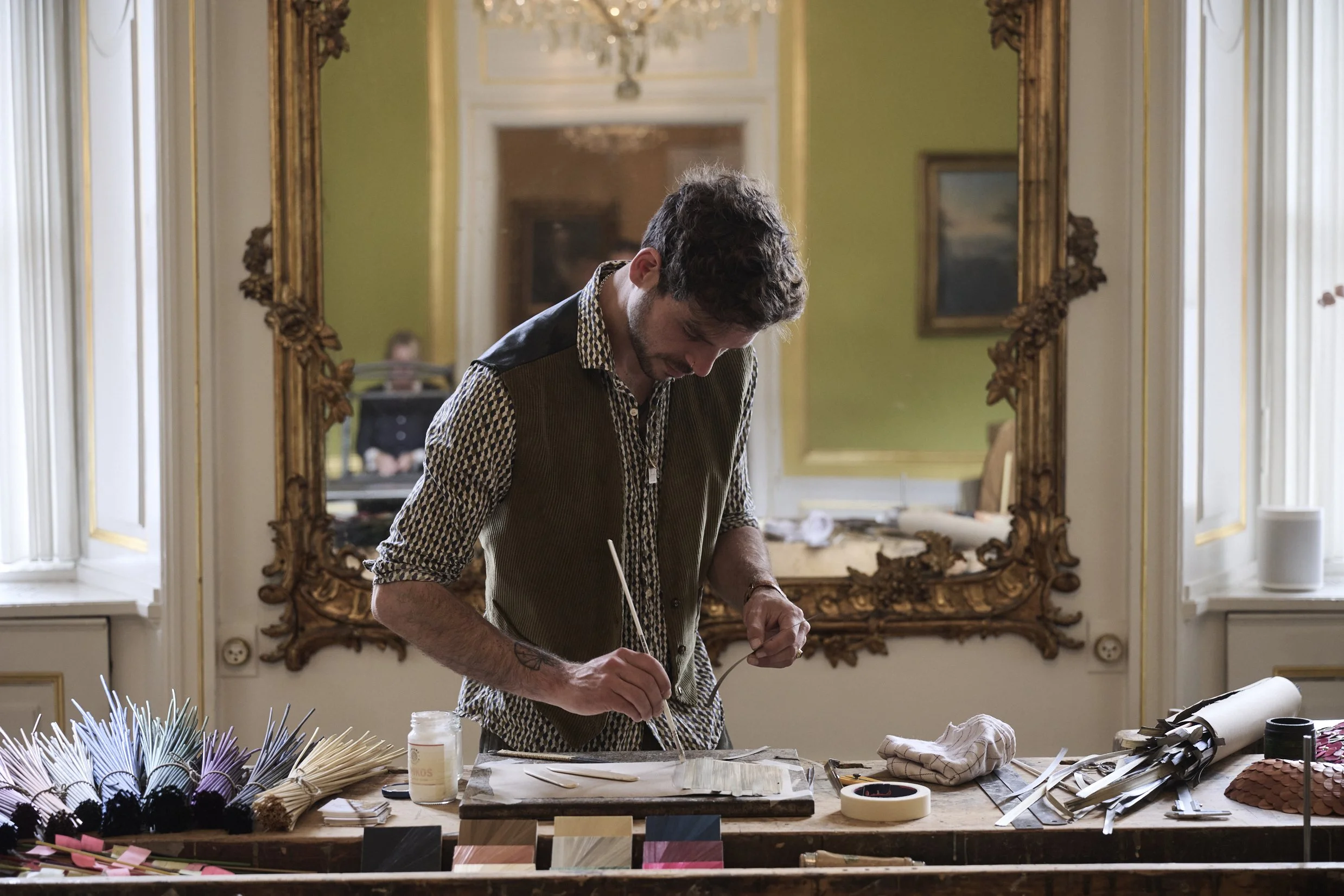 A man with curly hair and a checked shirt working on a craft project at a large wooden table in a room with ornate gold-framed mirror and paintings on green walls.