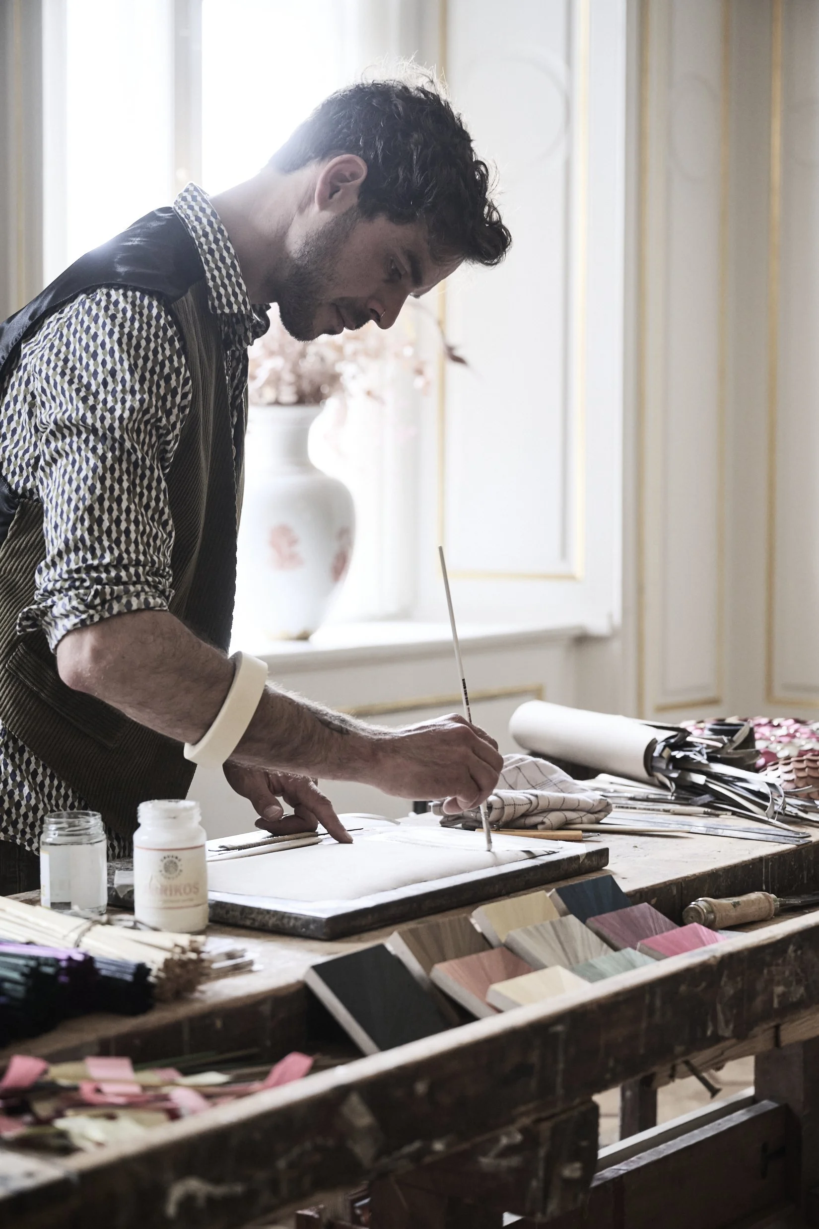 Jonathän Jouan working at a furniture workshop, painting or finishing a piece of furniture, surrounded by various supplies and color samples.