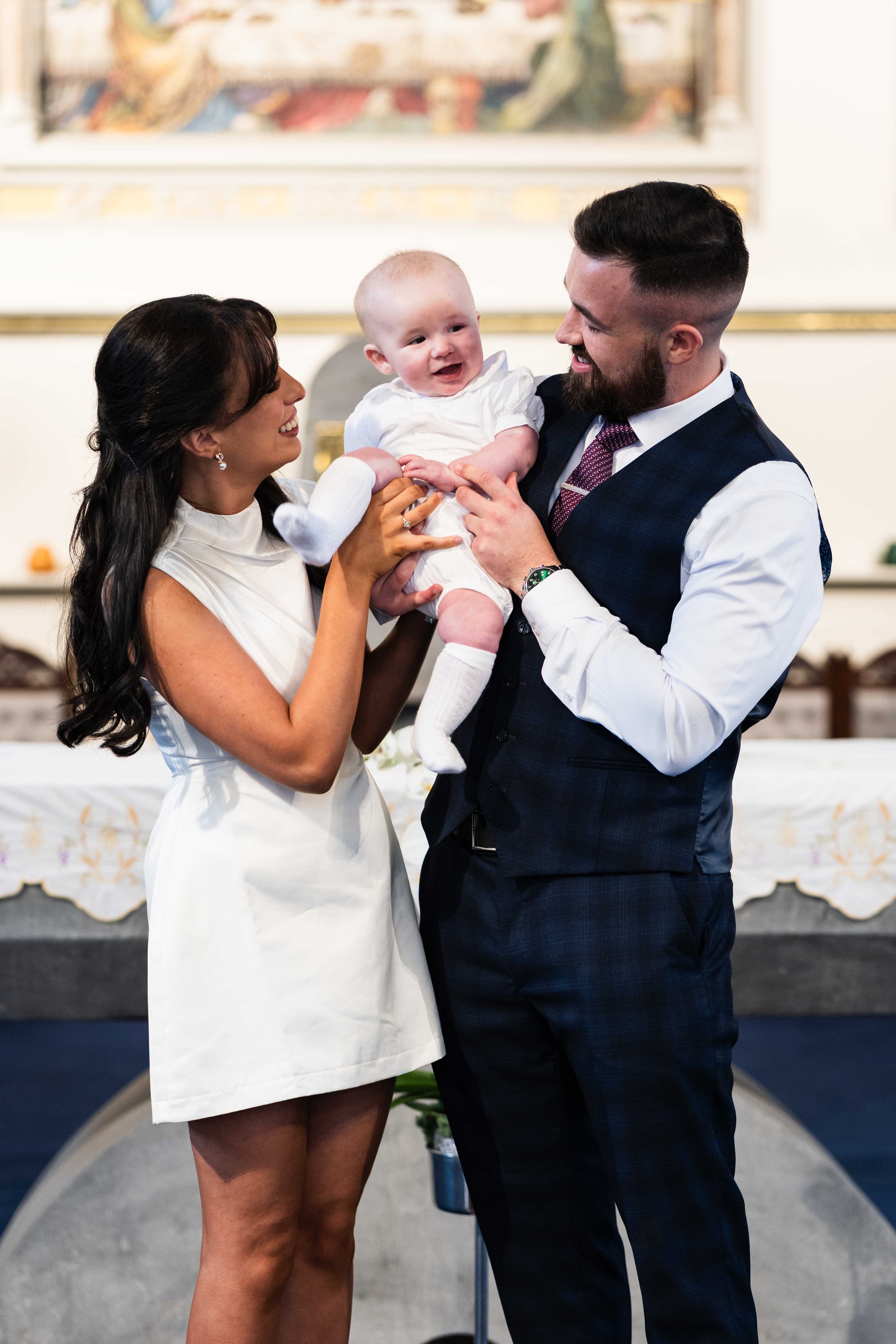 A family during a religious ceremony, with a woman holding a baby boy, and a man smiling at them. Christening photography Ireland