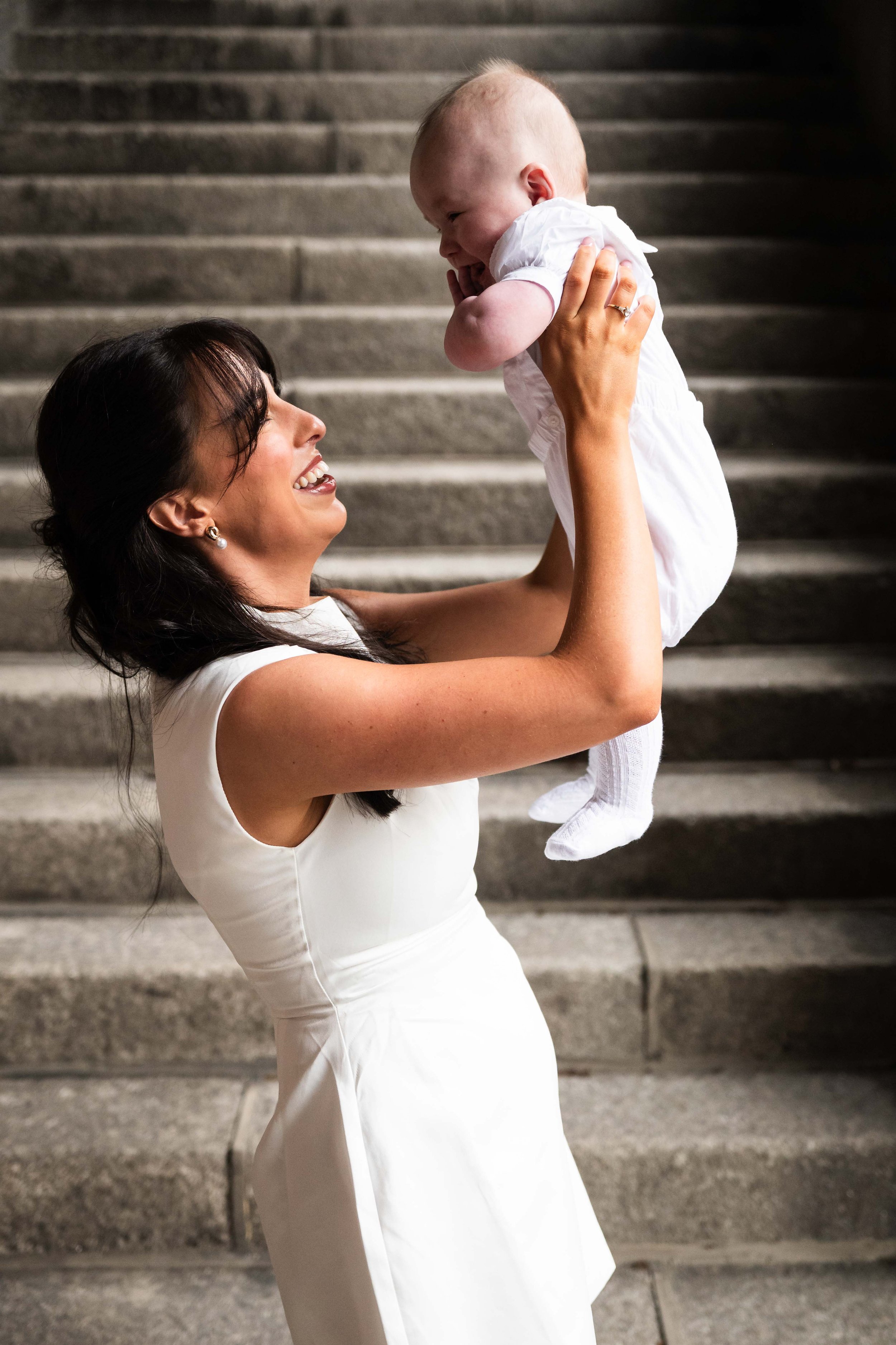 A woman in a white dress lifting a smiling baby in white clothing on a staircase. Christening photography Ireland