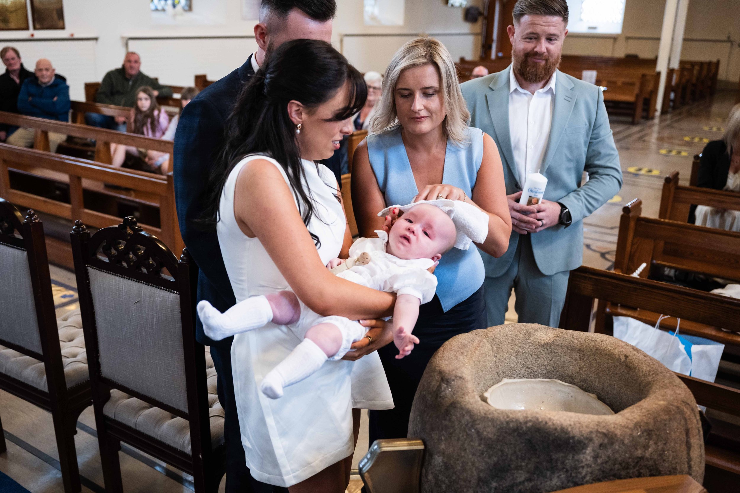 A woman holding a baby during a baptism ceremony in a church, with other adults and children in the background. Christening photography Ireland