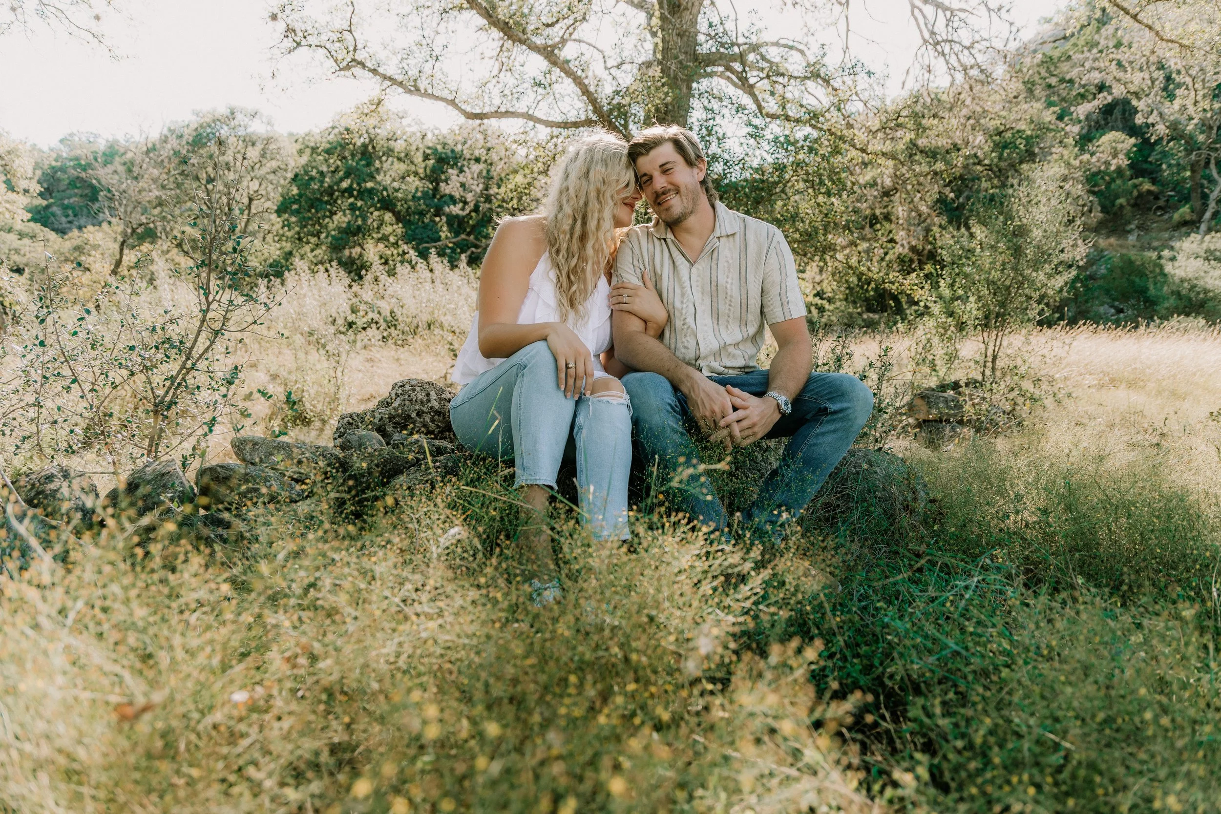 A couple sitting on rocks in a field with trees and bushes in the background, smiling and leaning close to each other.
