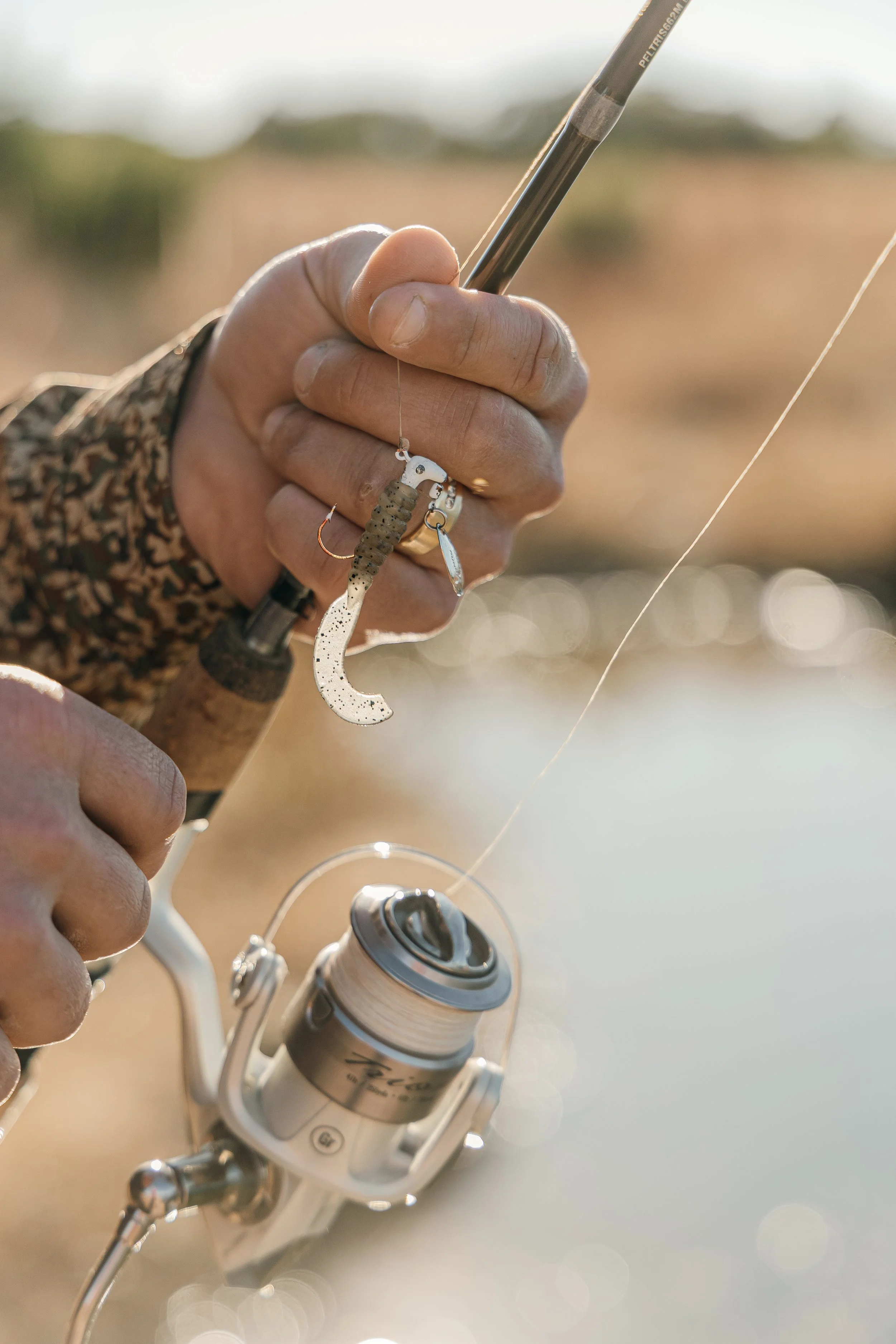 Close-up of a person's hand holding a fishing rod with a lure, fishing near a body of water during daytime.