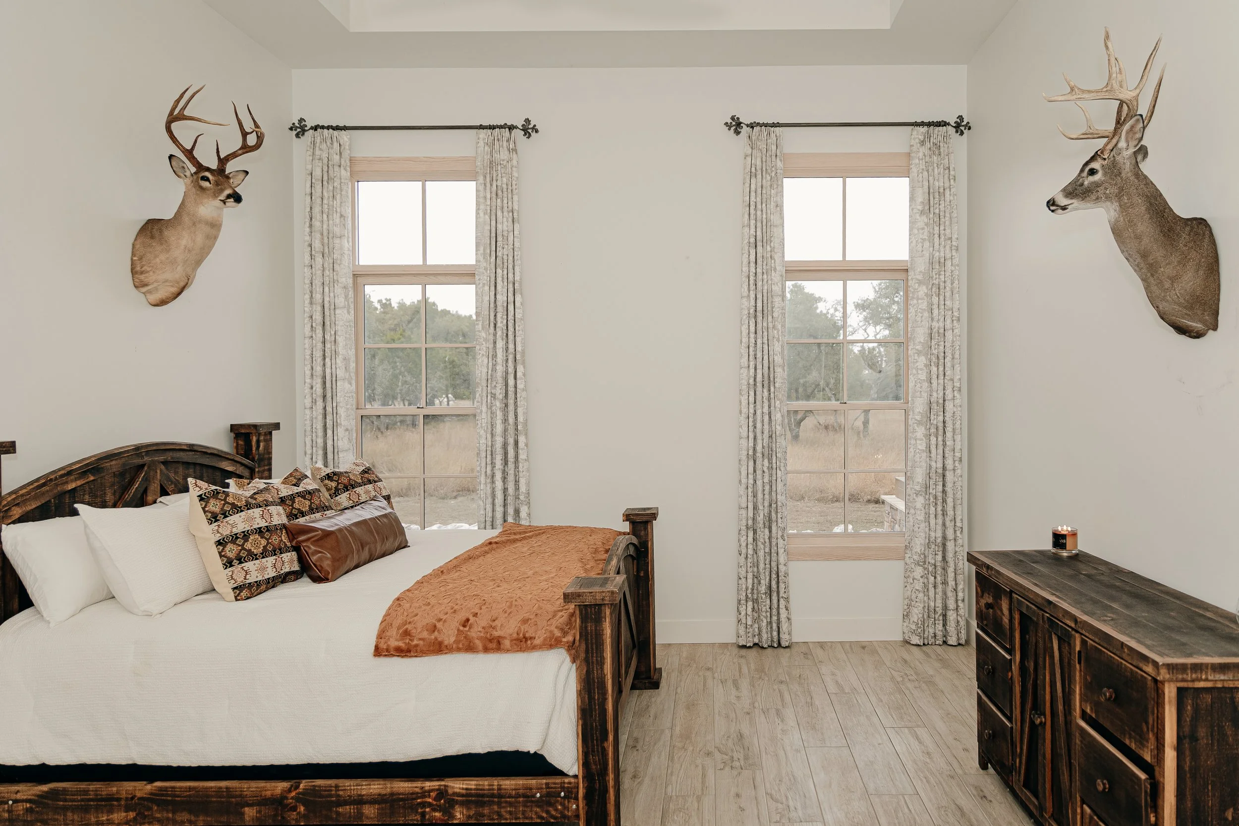 Bedroom with a wooden bed, white bedding, pillows, and a rust-colored blanket. Two windows with floral curtains, and mounted deer heads on the walls.