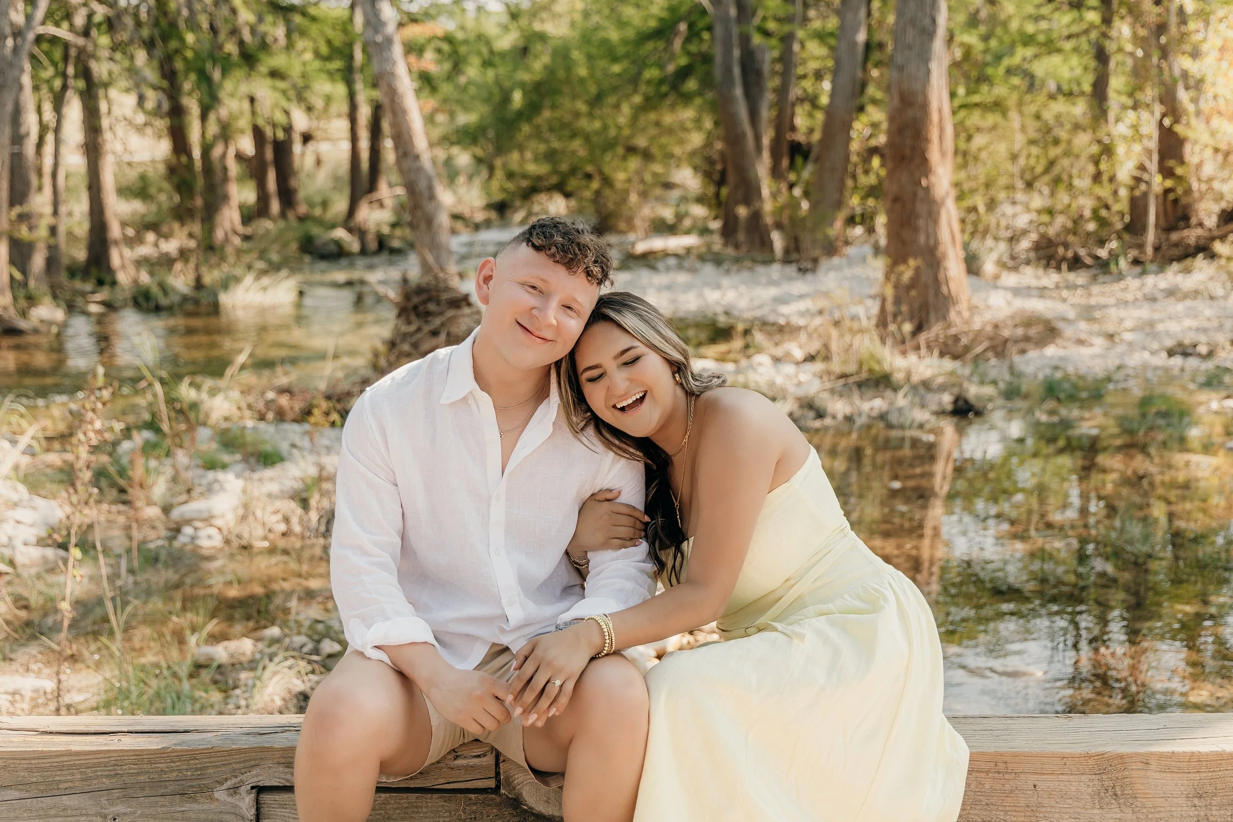 A happy couple sitting on a wooden log by a creek in a forest, smiling and laughing, with trees and water in the background.