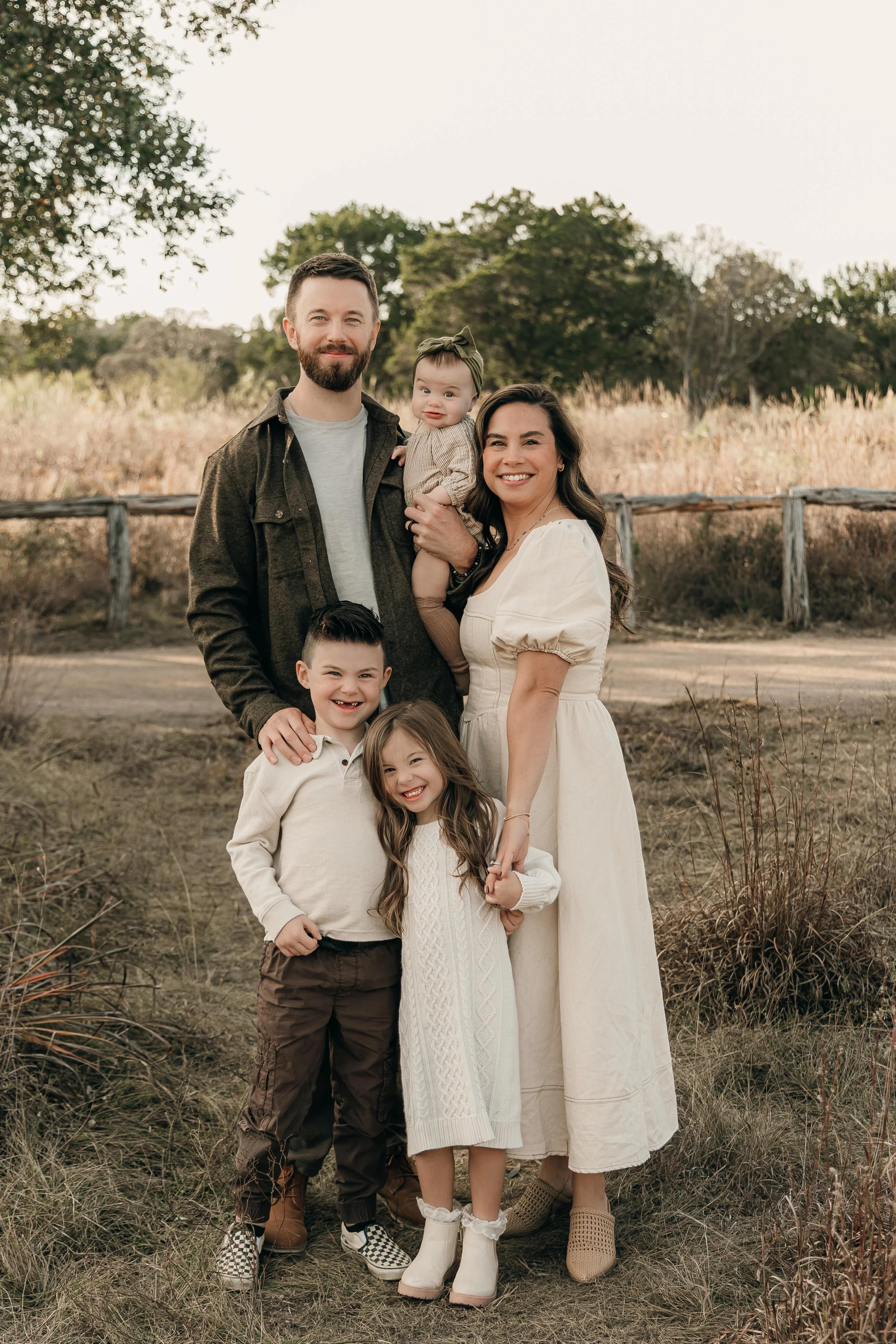 A family of six outdoors, standing in a field with grass and trees in the background, smiling at the camera.
