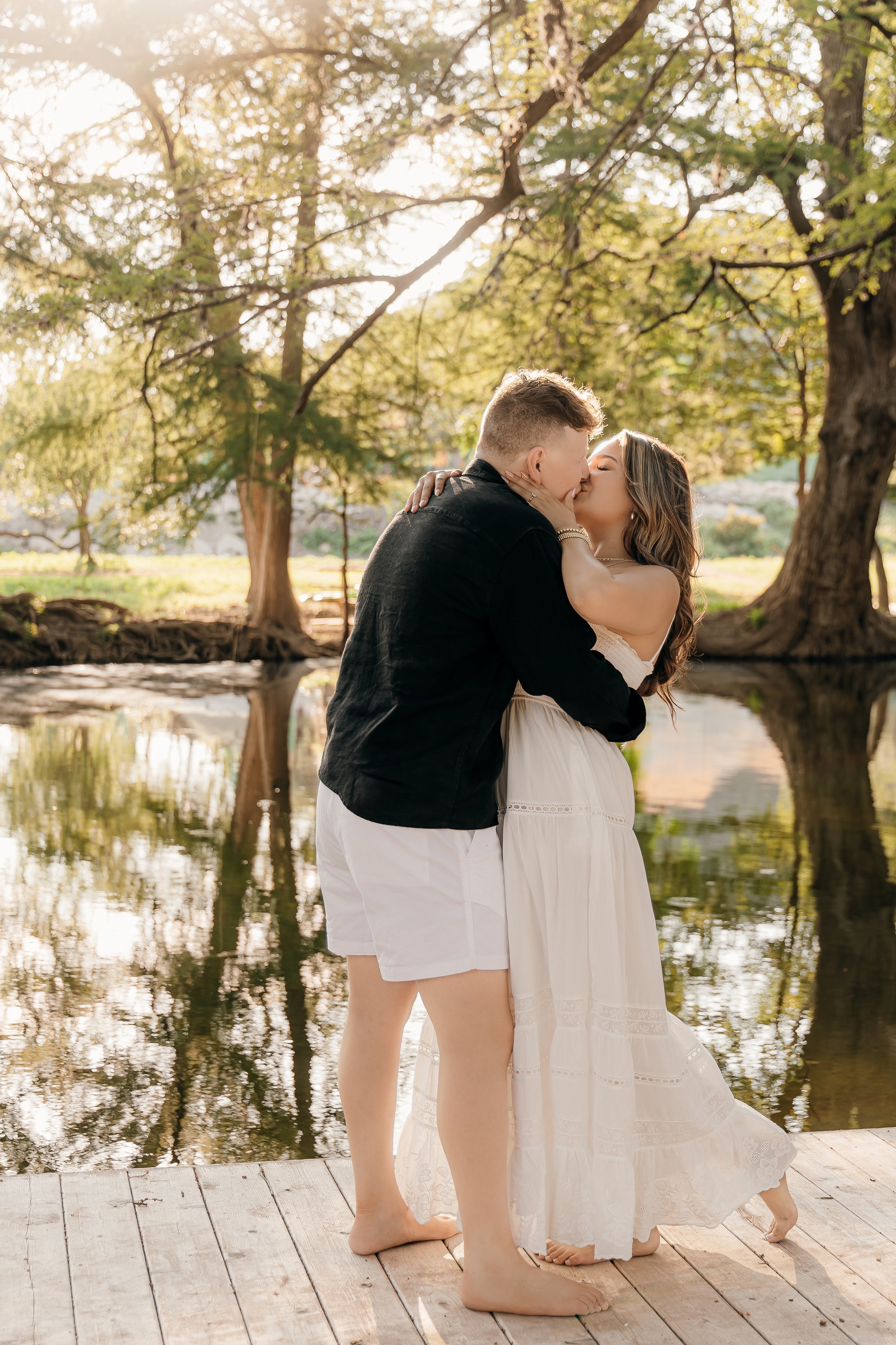A couple sharing a kiss by a pond in a park during sunset, surrounded by trees with green leaves.