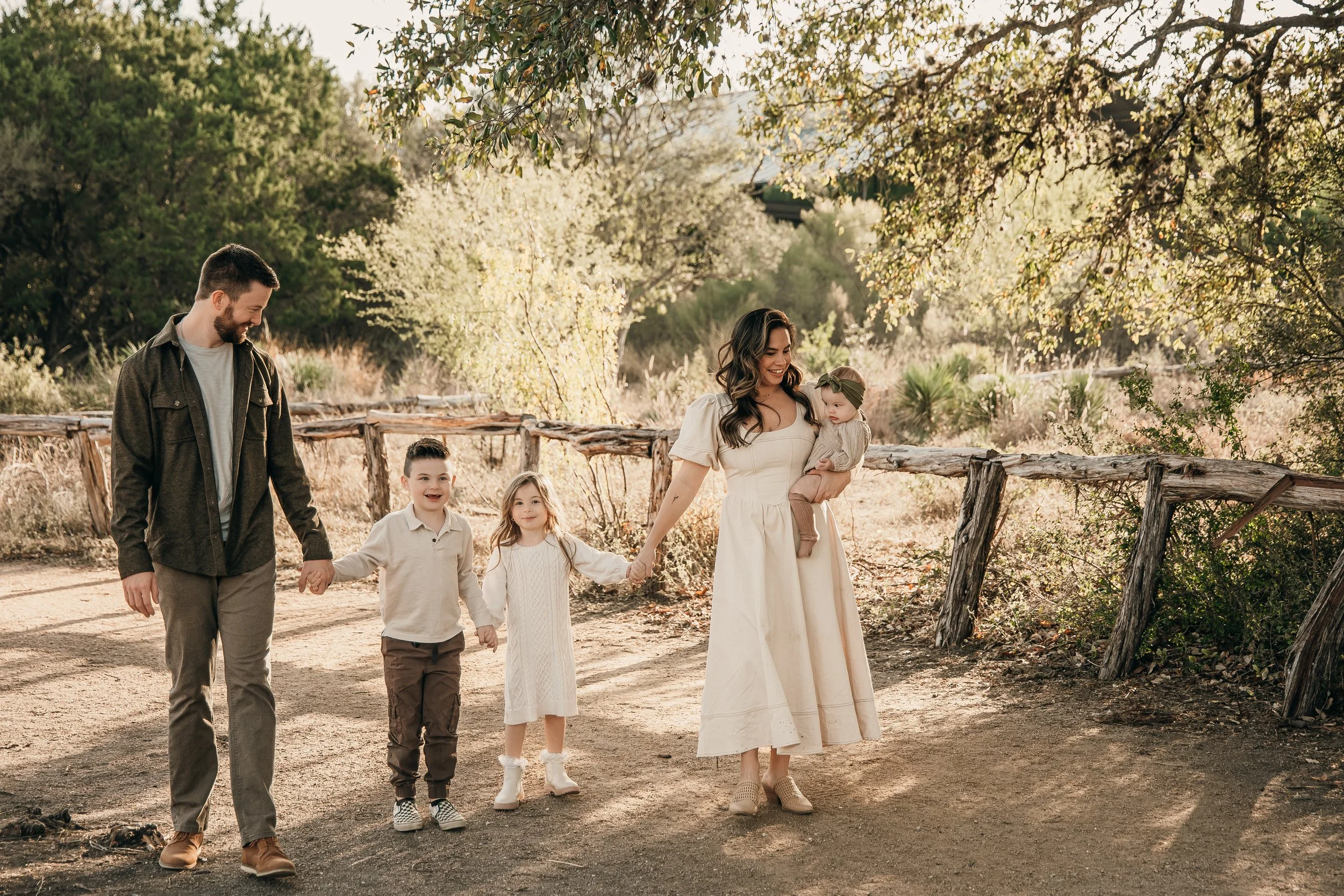 A family of five walking outdoors on a dirt path, holding hands and smiling, surrounded by trees and greenery during daytime.