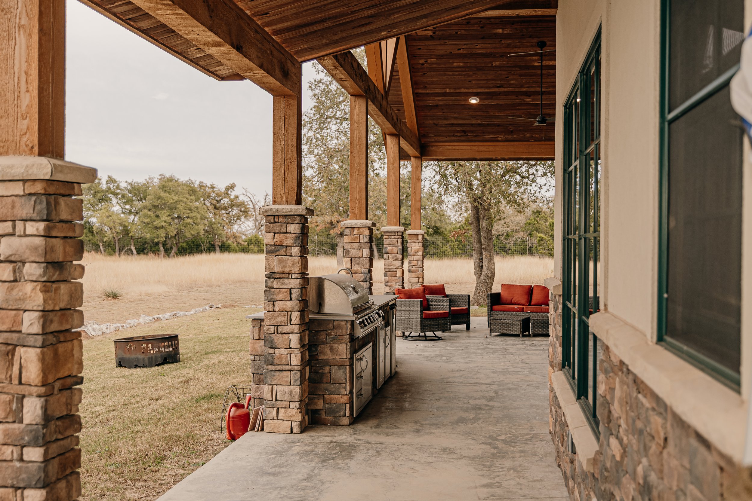 Covered outdoor patio with stone columns, wooden ceiling, outdoor grill, and seating area with orange cushions amidst a grassy field with trees in the background.