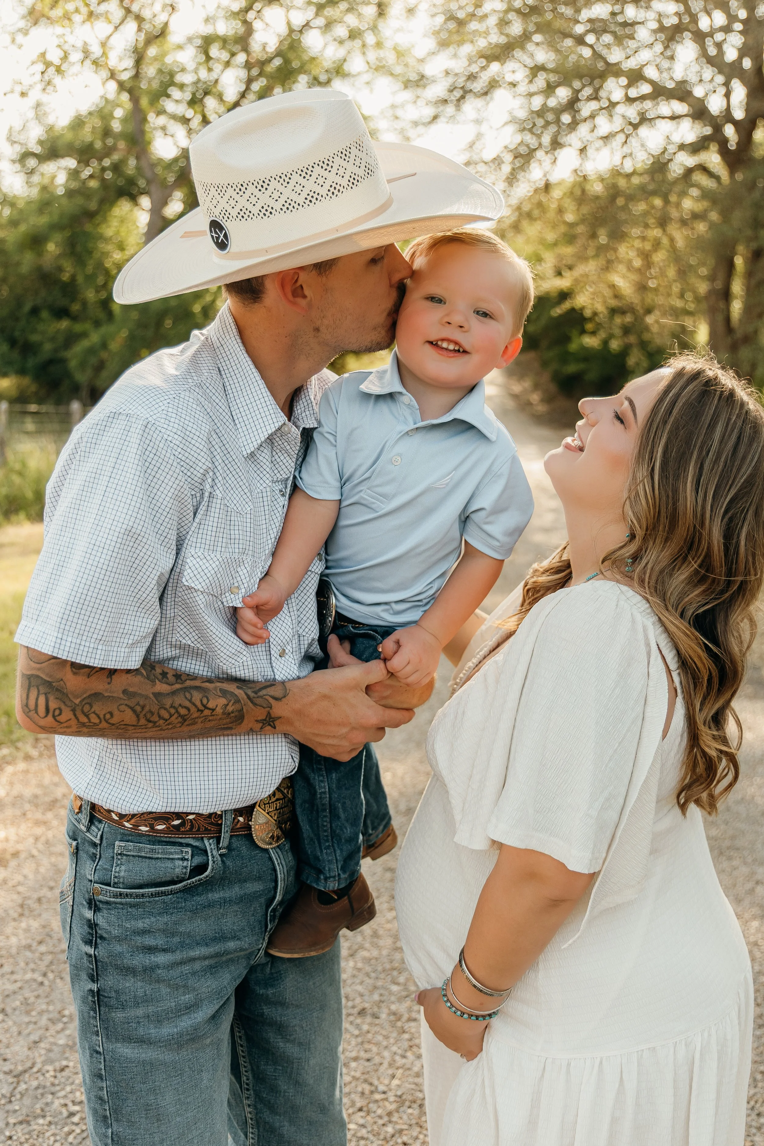 A family of three outdoors during the late afternoon. The father, wearing a cowboy hat and casual clothes, is kissing his young son on the cheek. The son is smiling, dressed in a light blue shirt. The mother, with long wavy hair and dressed in a whit