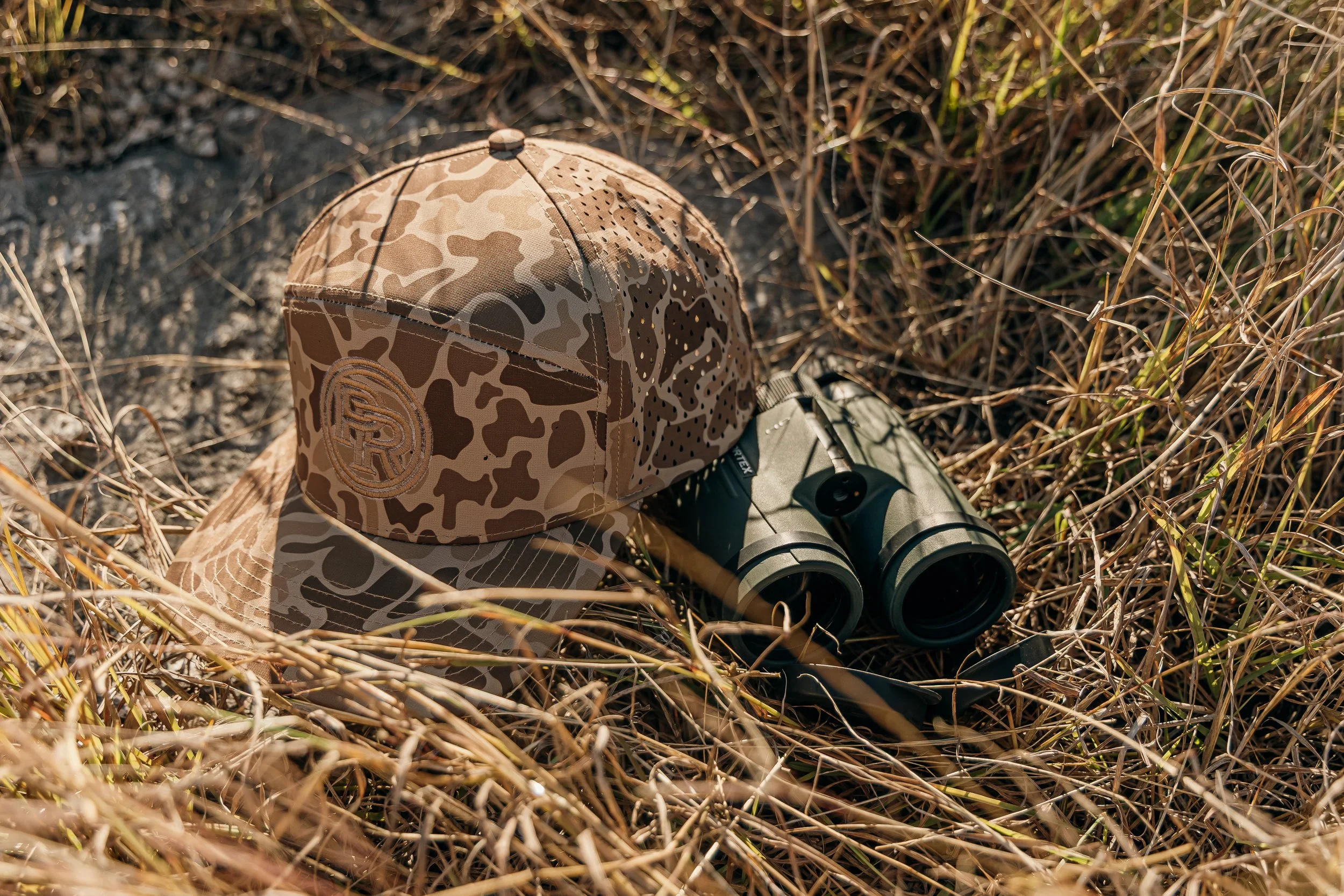 A camouflage baseball cap and black binoculars lying on dry grass in an outdoor setting.