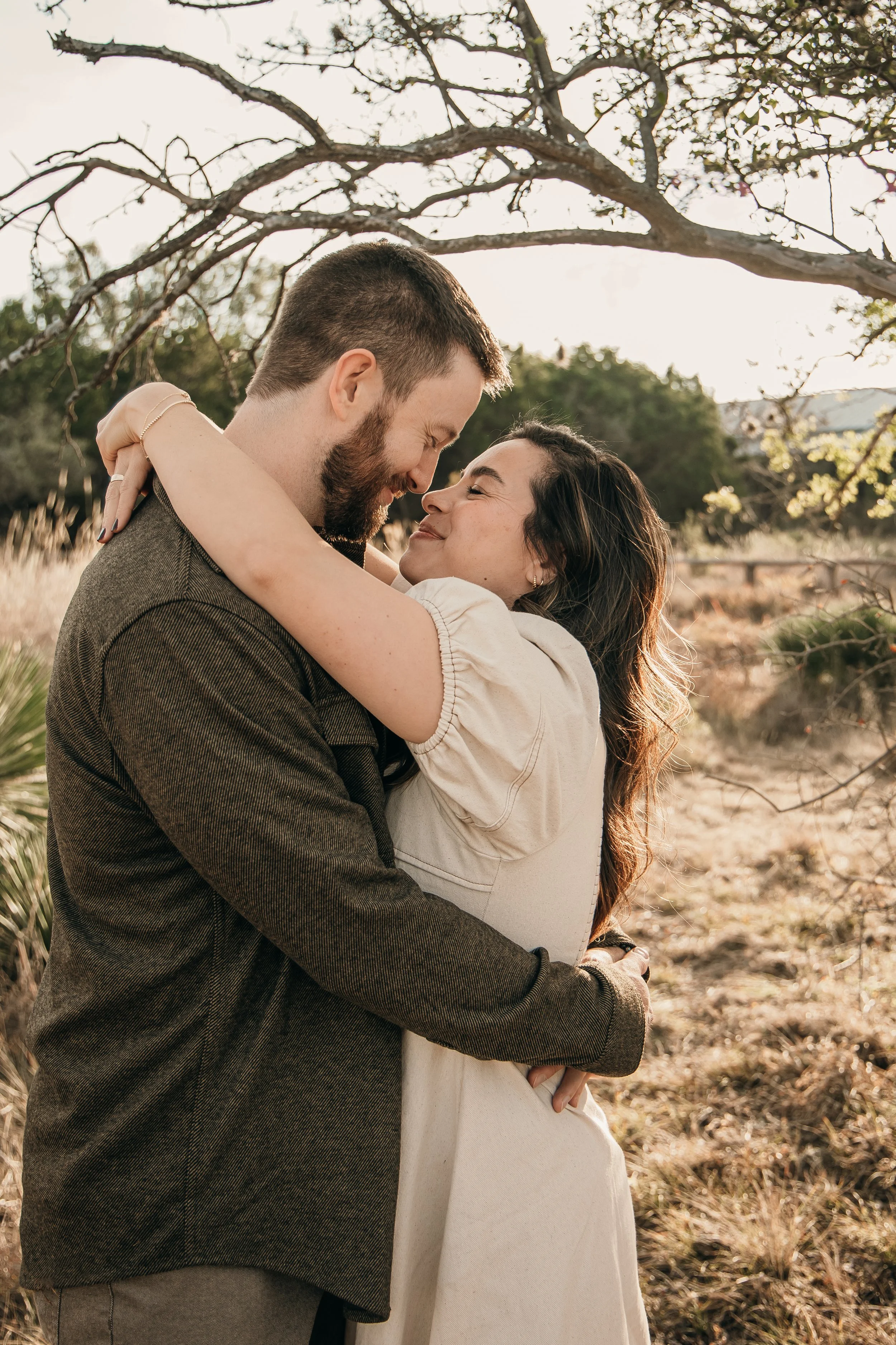 A couple embracing outdoors under a tree, sharing a joyful and intimate moment, with the man wearing a brown jacket and the woman in a light-colored dress.