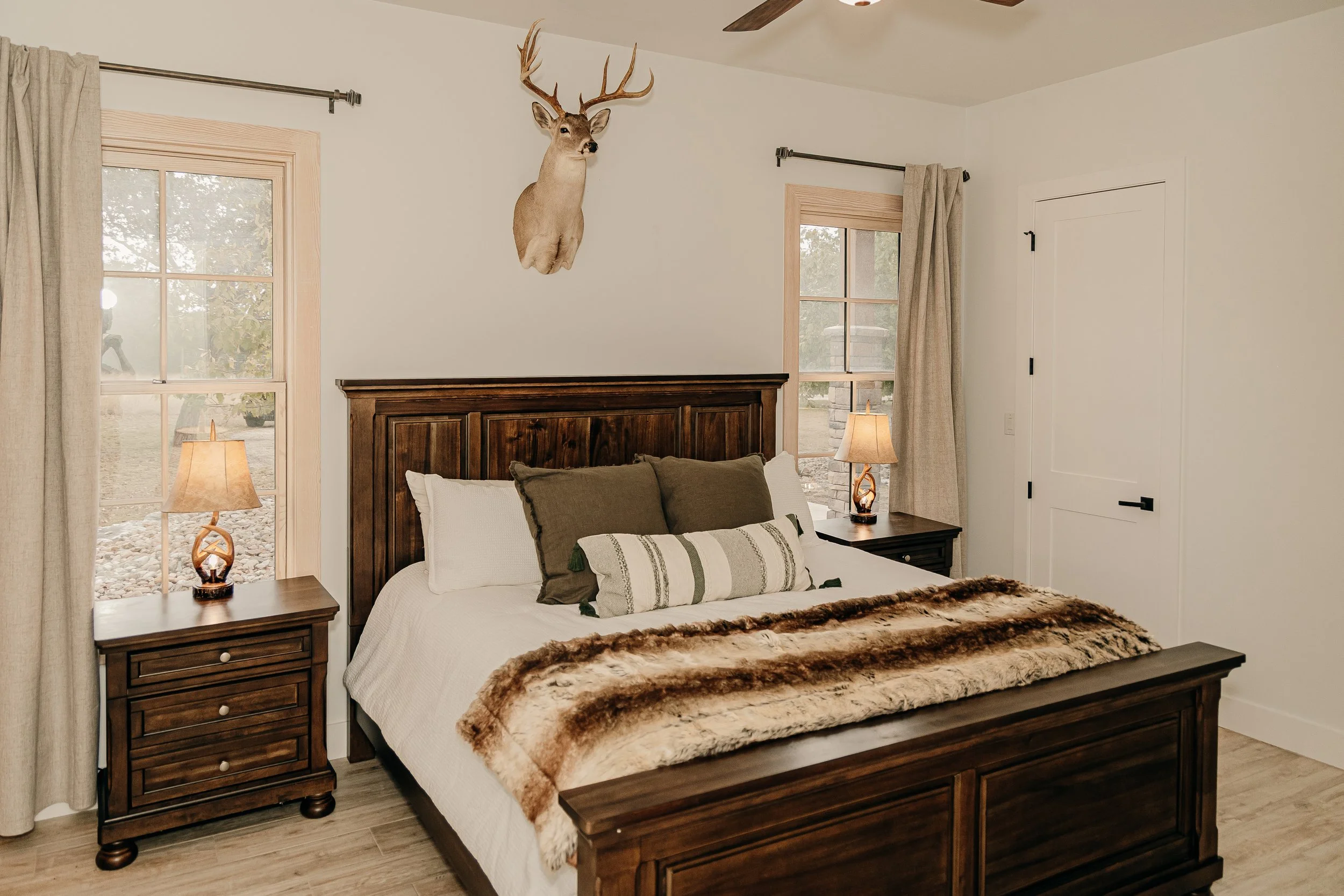 A bedroom with a large wooden bed, a deer head mounted on the wall above, two bedside tables with lamps, two windows with beige curtains, and a ceiling fan.