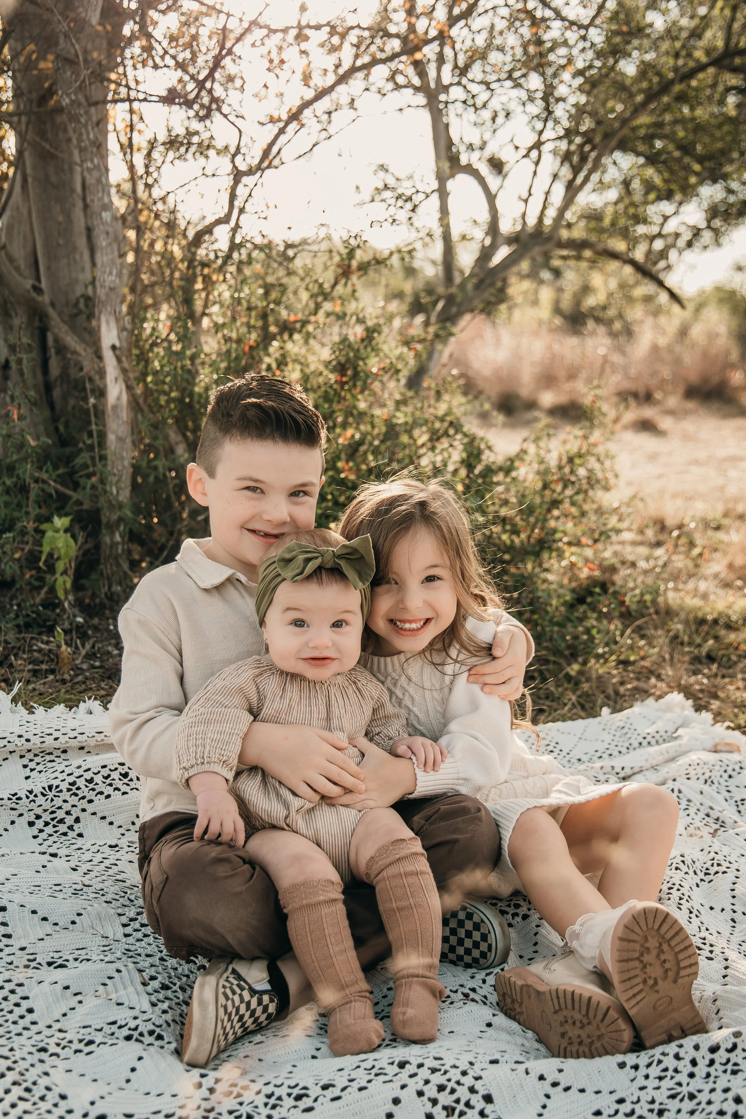 Three children, two girls and one boy, sitting on a white crocheted blanket outdoors, hugging and smiling at the camera, with trees and sunlight in the background.
