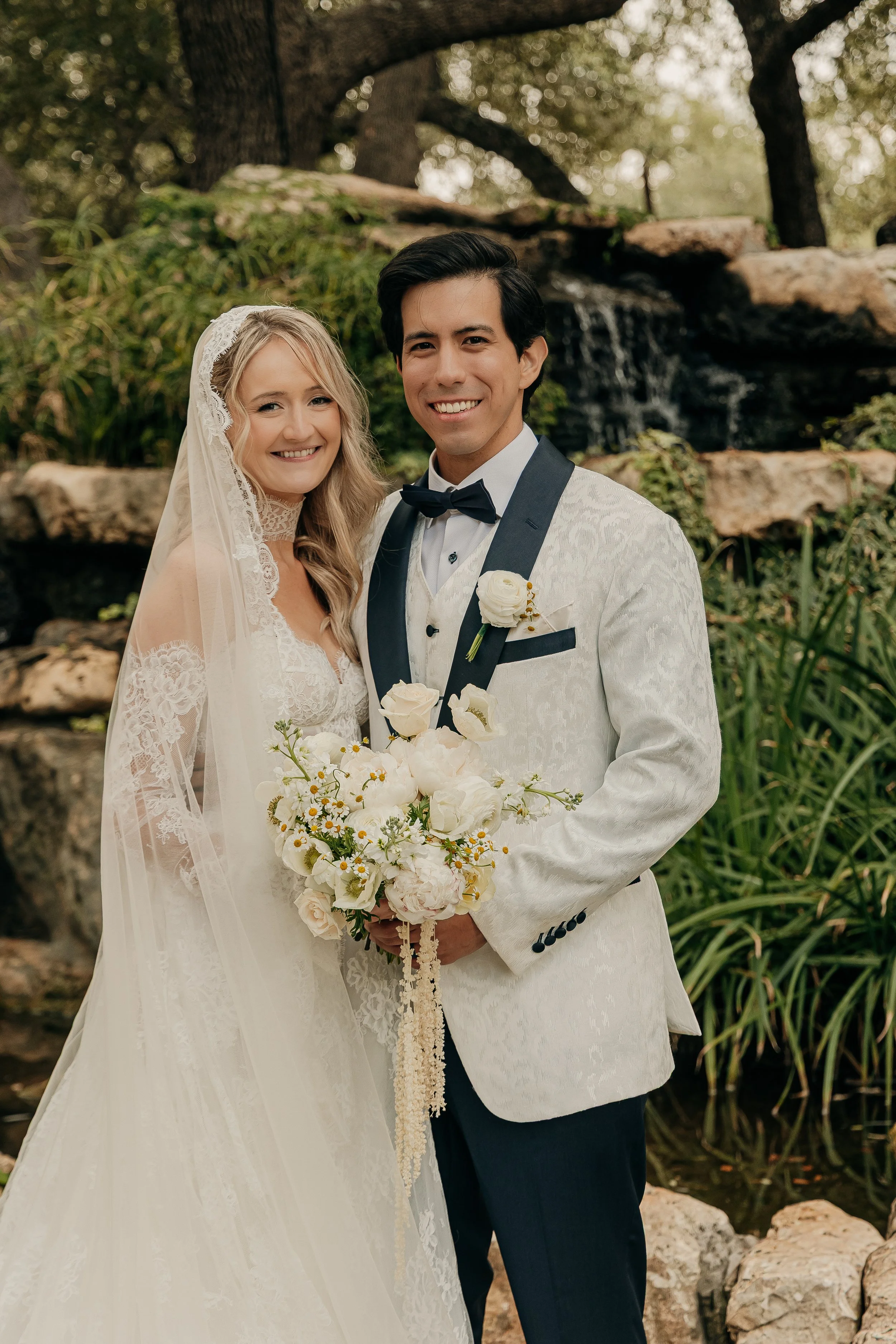 A newlywed couple in wedding attire stands outdoors by a waterfall, smiling at the camera. The bride is wearing a lace wedding dress with a veil, and the groom is dressed in a white tuxedo with a black bow tie, holding a bouquet of white flowers.