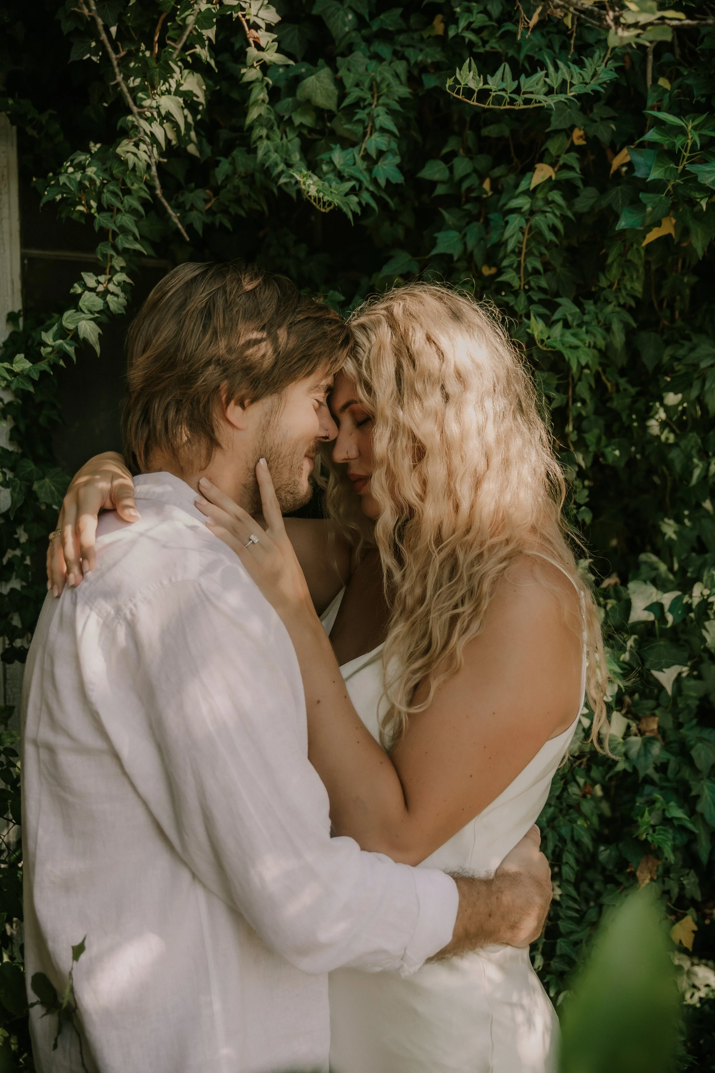 A couple embracing closely with eyes closed, touching foreheads, in front of green ivy leaves.