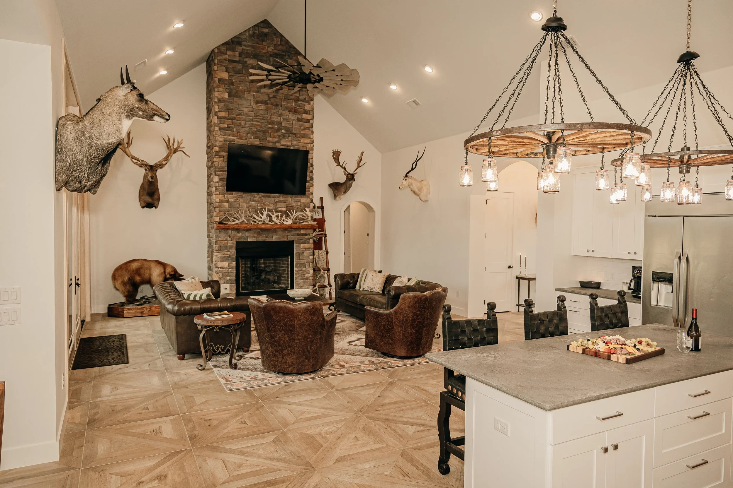 Living room with a vaulted ceiling, a brick fireplace, mounted animal heads, a mounted bear, a TV above the fireplace, leather and upholstered chairs, and rustic hunting decor. An open kitchen with a countertop and dining area visible in the foregrou