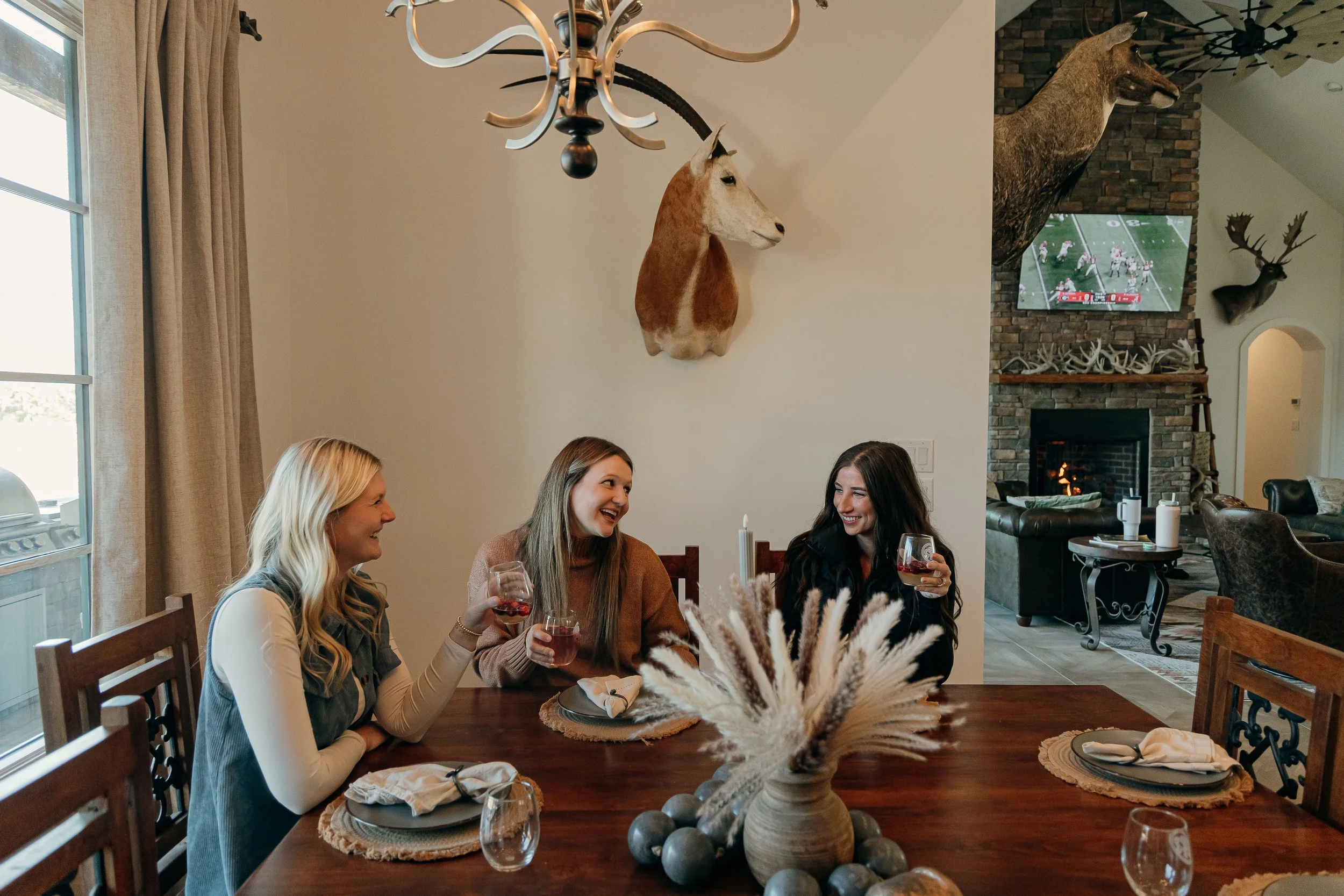 Three women sitting at a dining table, smiling and holding glasses of red wine, in a cozy home with deer and mountain goat taxidermy on the wall, a fireplace, and a mounted deer head in the background.