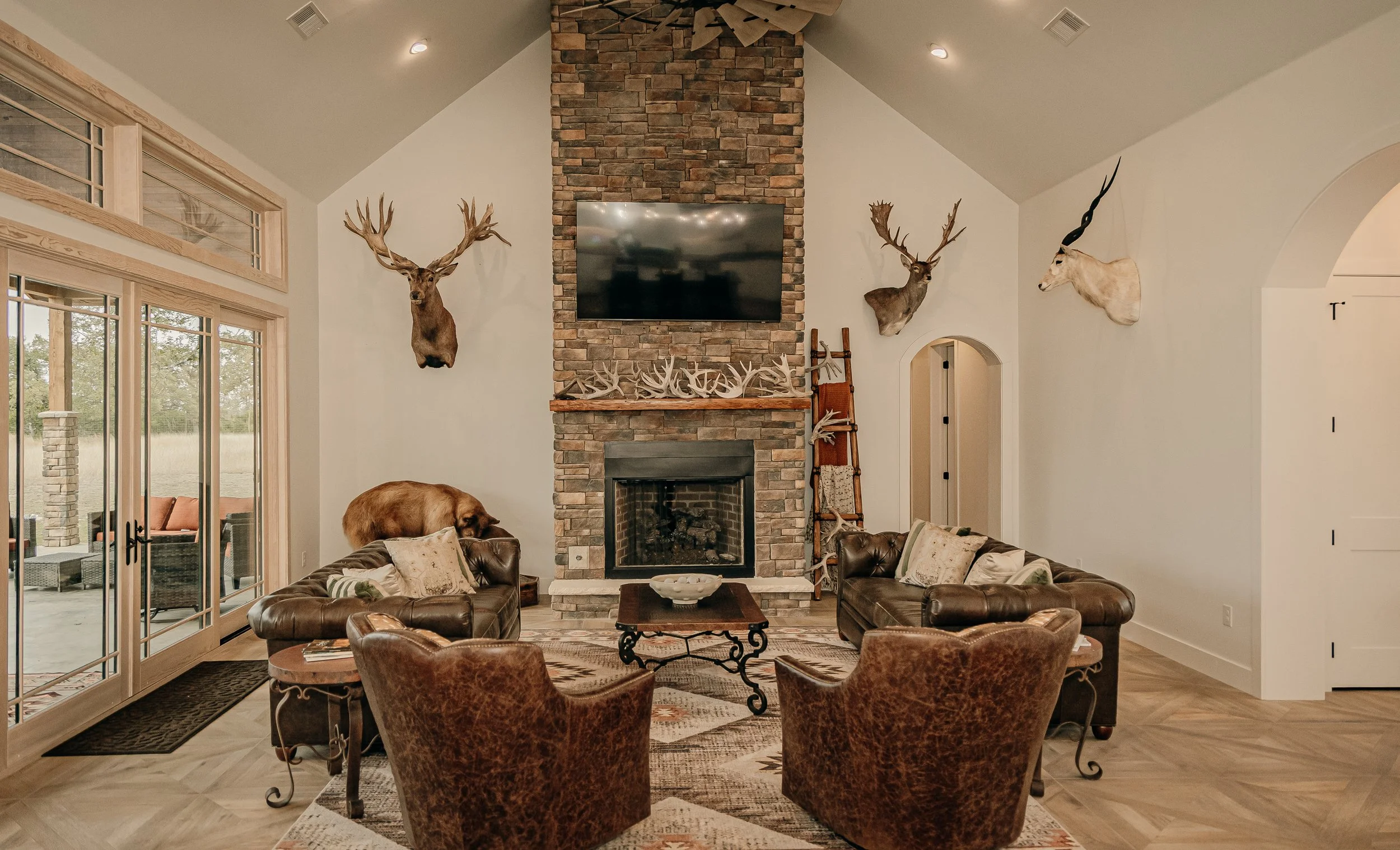 Living room with a brick fireplace, mounted television, and deer and antelope head mounts on the wall. Contains leather sofas, armchairs, and a wooden coffee table. A bear rests on the sofa near sliding glass doors that lead outside.