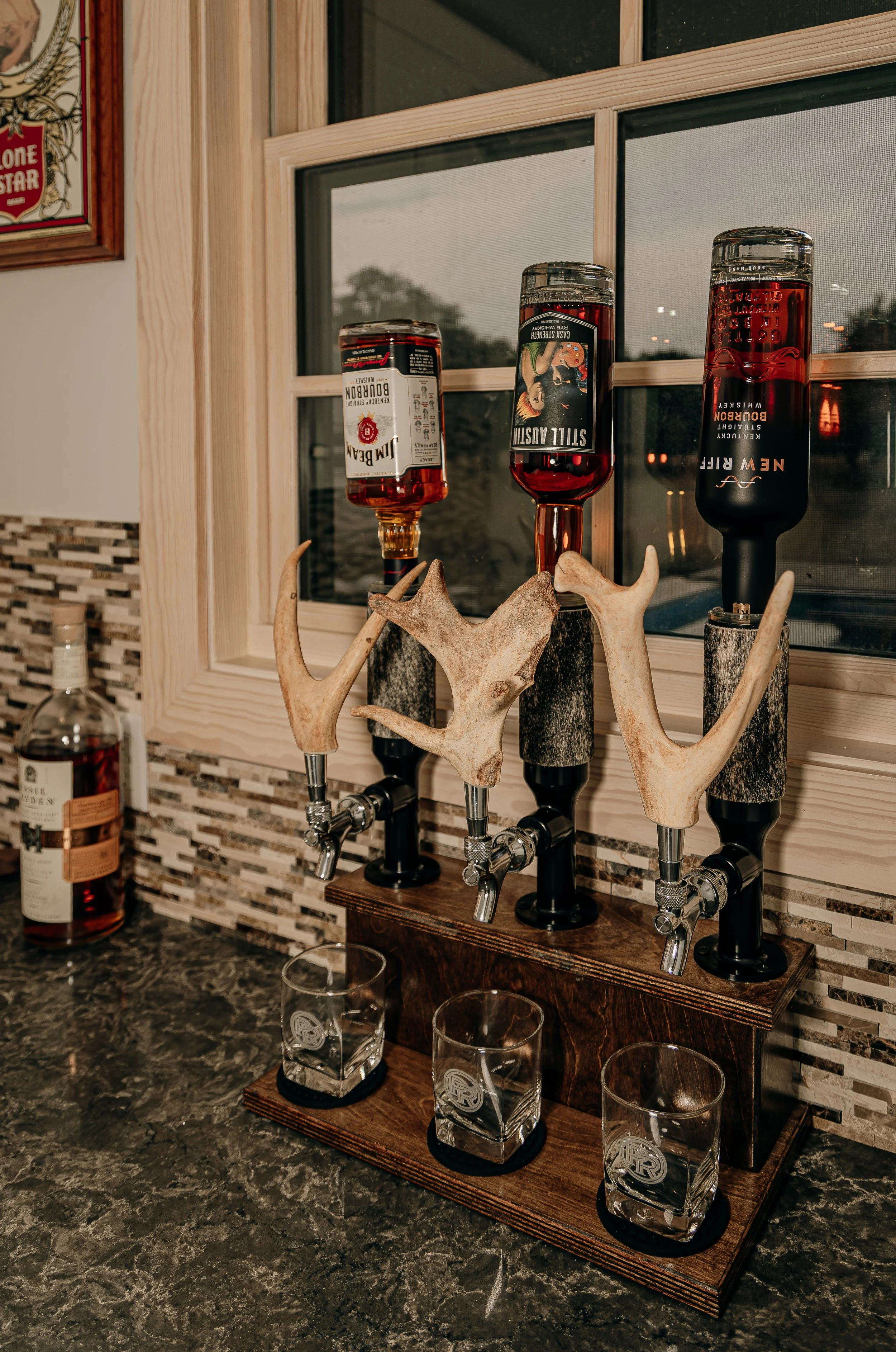 A decorative liquor dispenser featuring antler-style spouts holding bottles of alcohol, with three glasses placed beneath the spouts on a wooden tray.