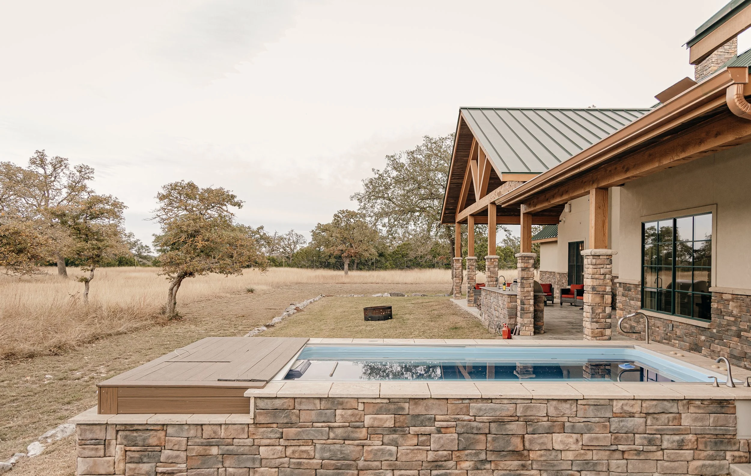Backyard view of a house with a covered porch, stone columns, and a nearby swimming pool with a cover, surrounded by dry grass and trees.