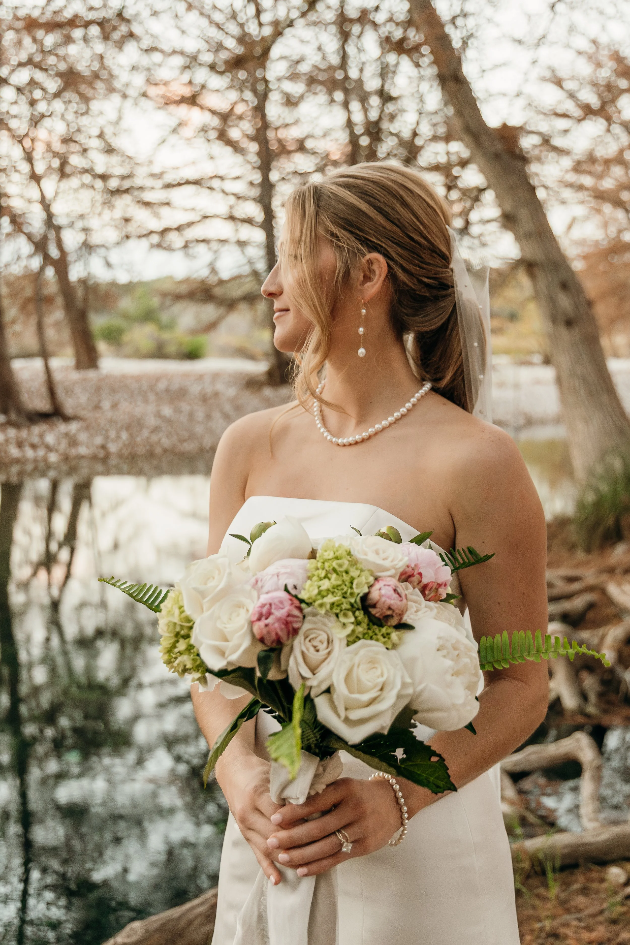 A bride in a strapless wedding dress holding a bouquet of white and pink flowers, standing near a pond in a wooded area with autumn leaves.
