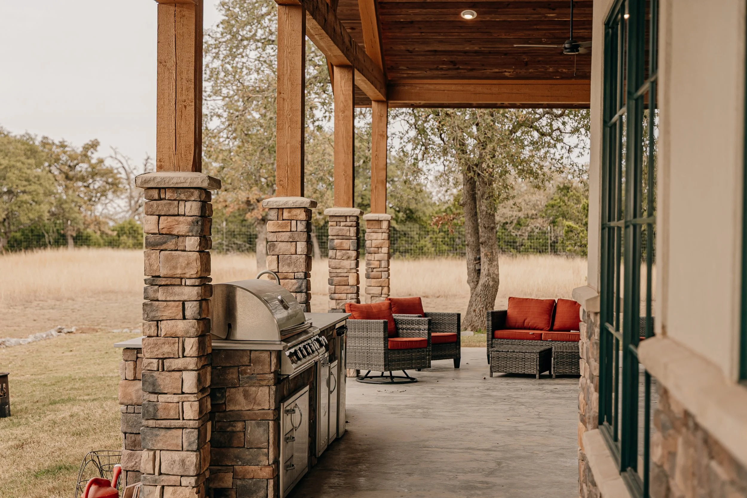 Covered outdoor patio with a built-in grill, stone columns, remaining outdoor furniture including a black wicker sofa set with red cushions, and trees in the background.