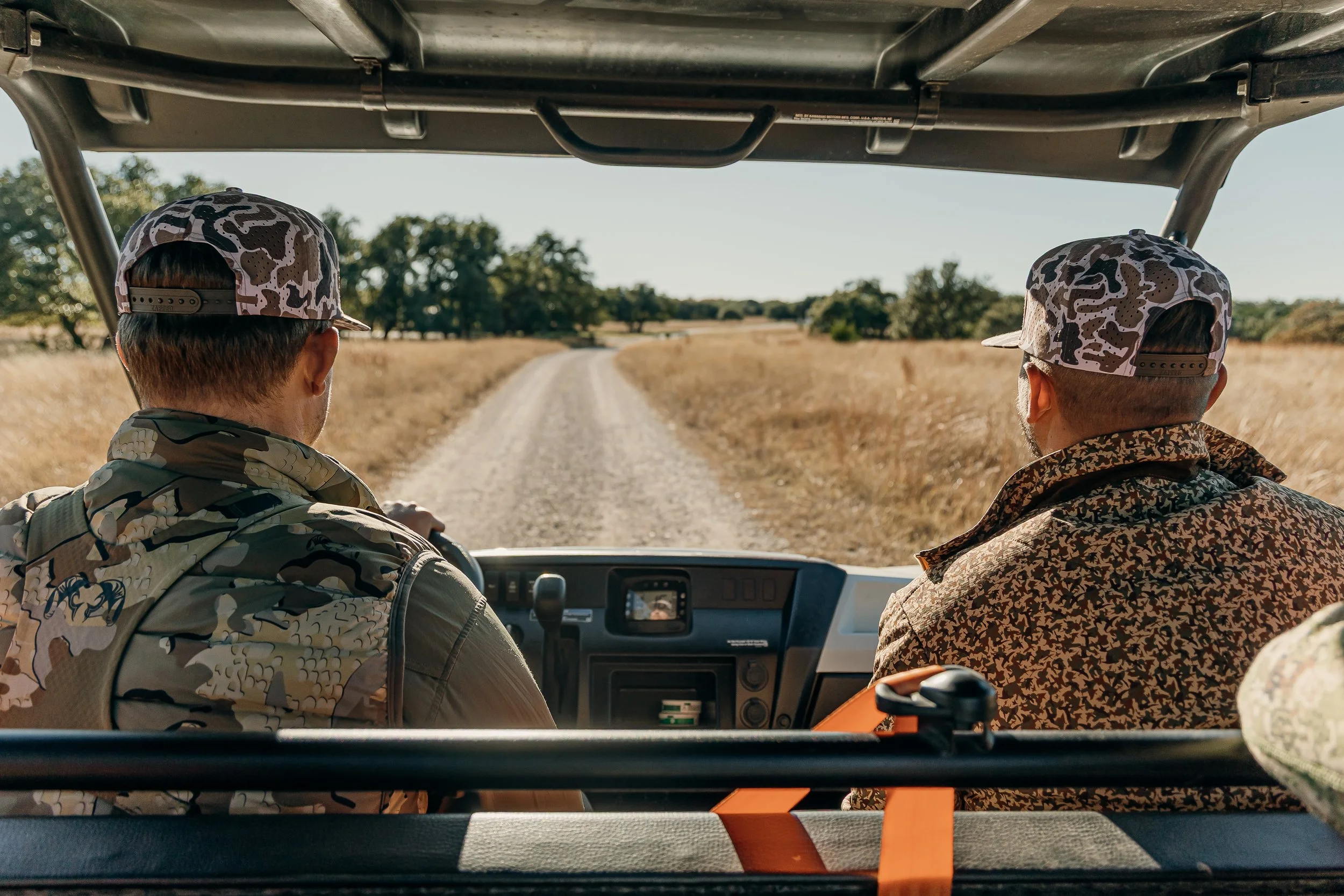 Two men riding in an open-air vehicle on a dirt road through a grassy field, wearing camouflage clothing and patterned baseball caps, with trees in the background.
