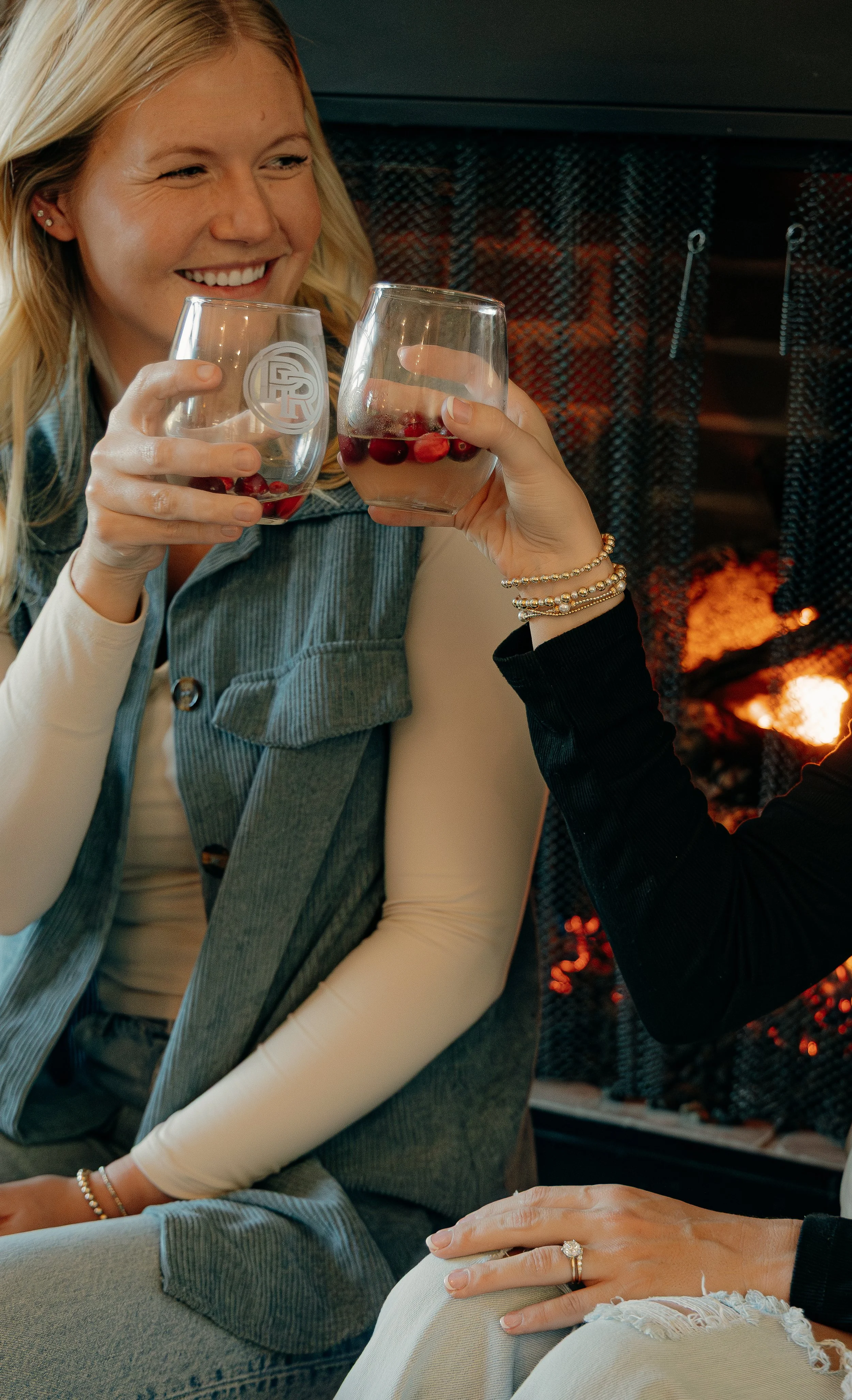 Two women clinking glasses filled with a beverage containing cherries in front of a fireplace, one smiling and wearing a denim vest and long-sleeve shirt, the other wearing jewelry and a dark top.