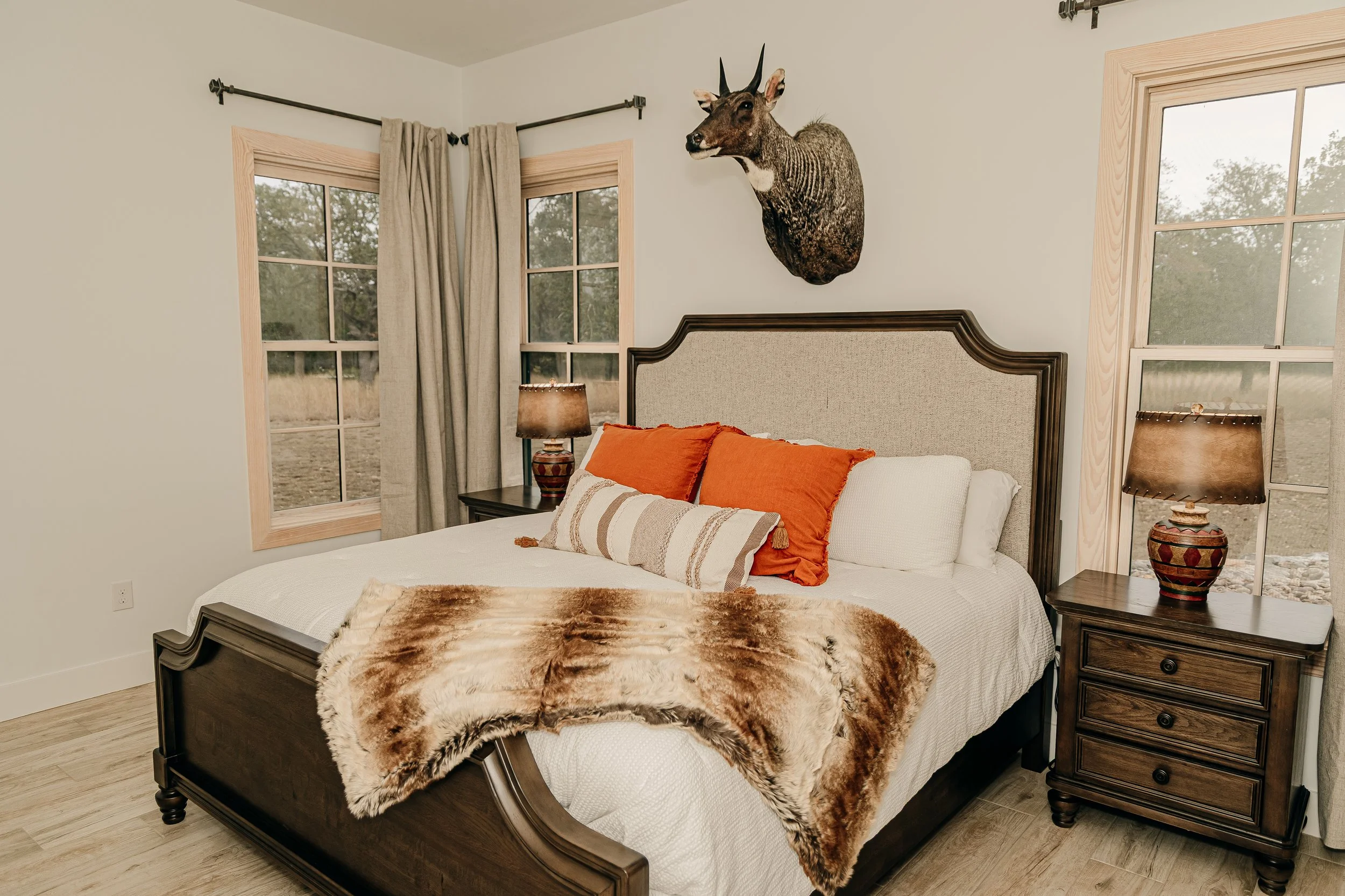 A bedroom with a large bed, two nightstands with lamps, windows with beige curtains, and a mounted deer head on the wall.