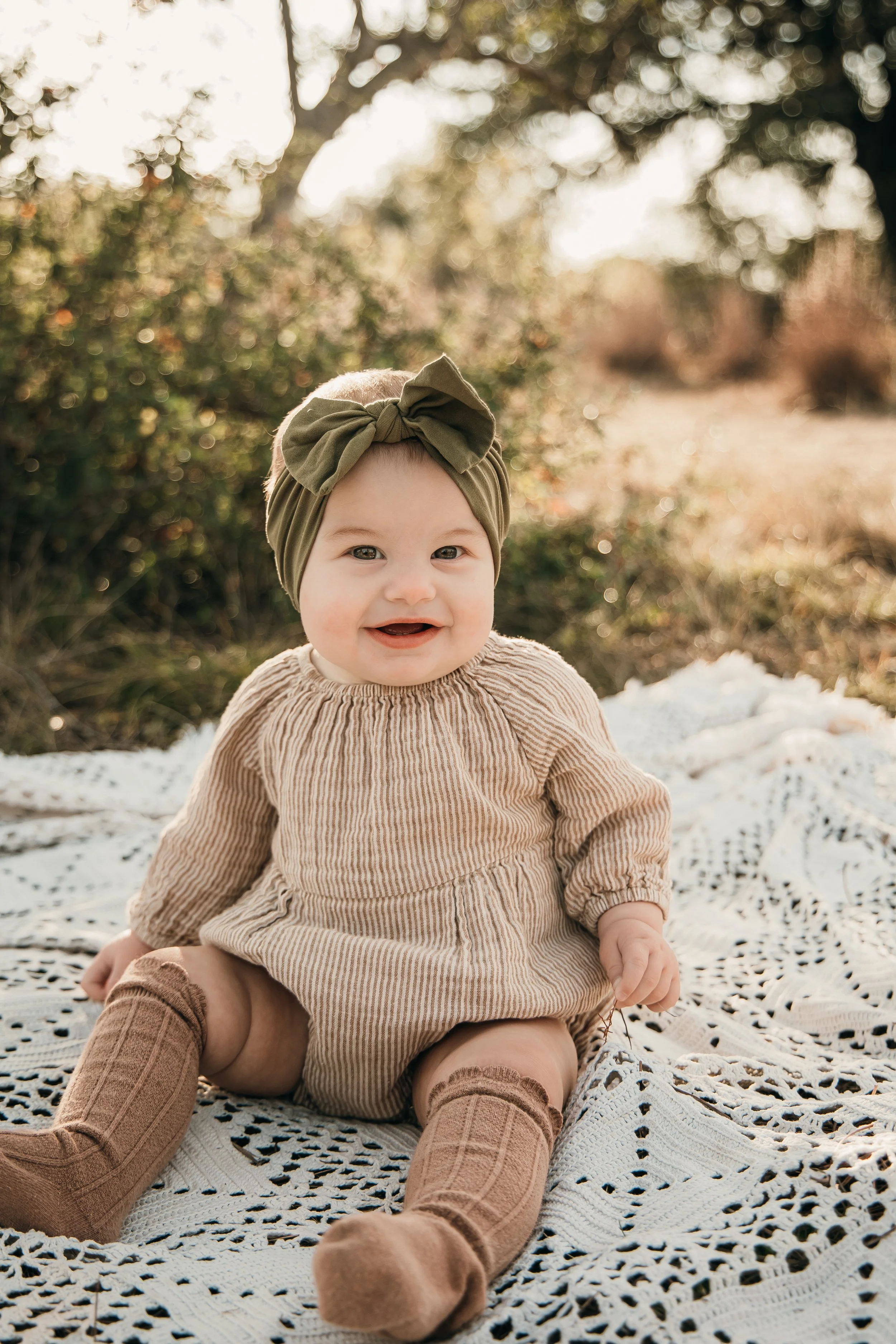 A smiling baby girl sitting outdoors on a white blanket, wearing a beige dress, brown knee-high socks, and a green headband with a bow. The background features trees and bushes with sunlight filtering through.